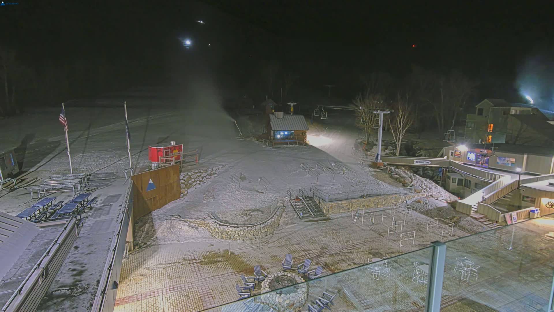 A snow-covered ski resort at night, illuminated by artificial lights, with snowmaking machines operating under a dark sky.
