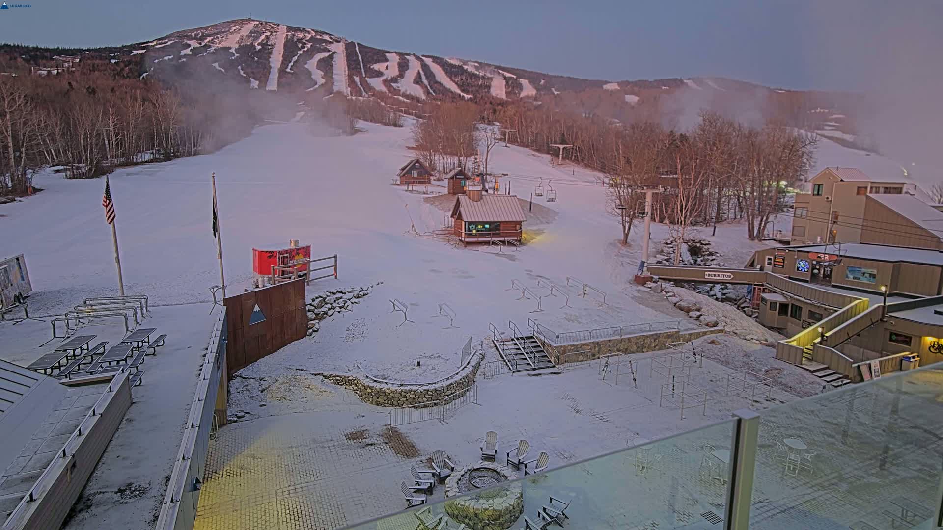 A snow-covered ski resort at dawn, with a mountain in the background and several buildings at the base.