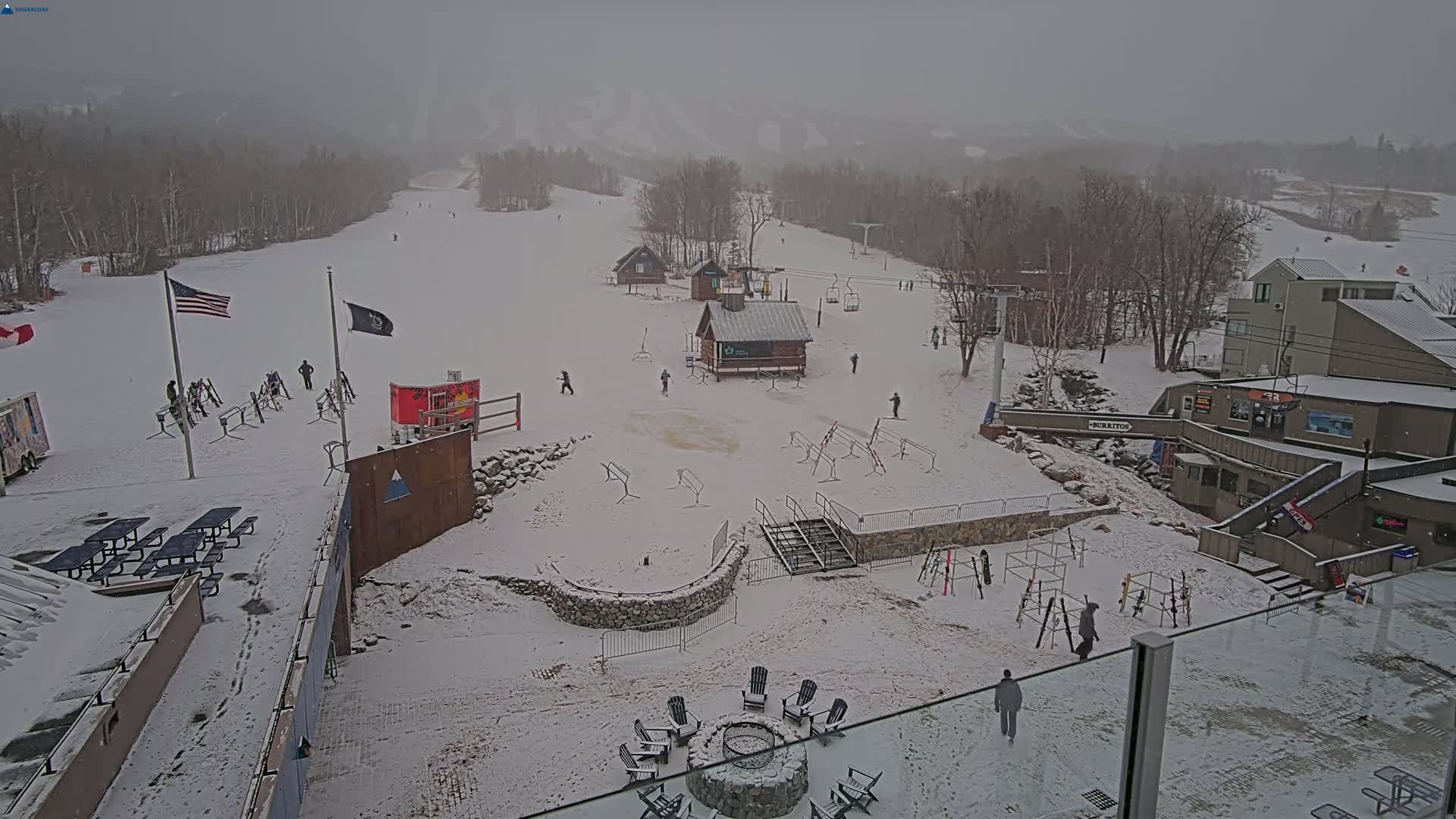 A snow-covered ski resort base area with several buildings, ski racks, picnic tables, and a few people, under an overcast sky.