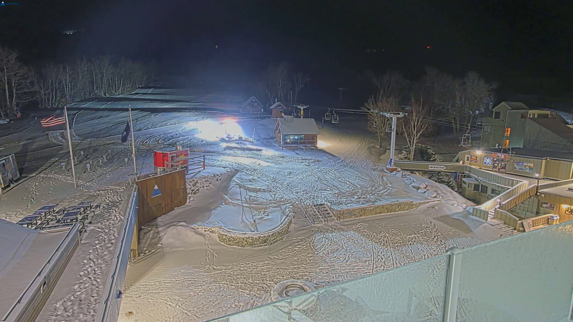 A snow-covered ski slope at night, illuminated by artificial lights, with buildings and ski lifts visible.