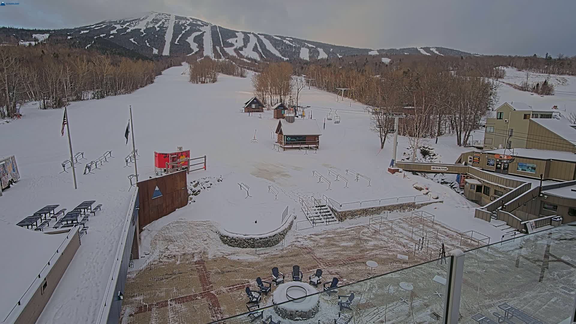 A snow-covered ski resort with several buildings, ski lifts, and seating areas is visible under an overcast sky.