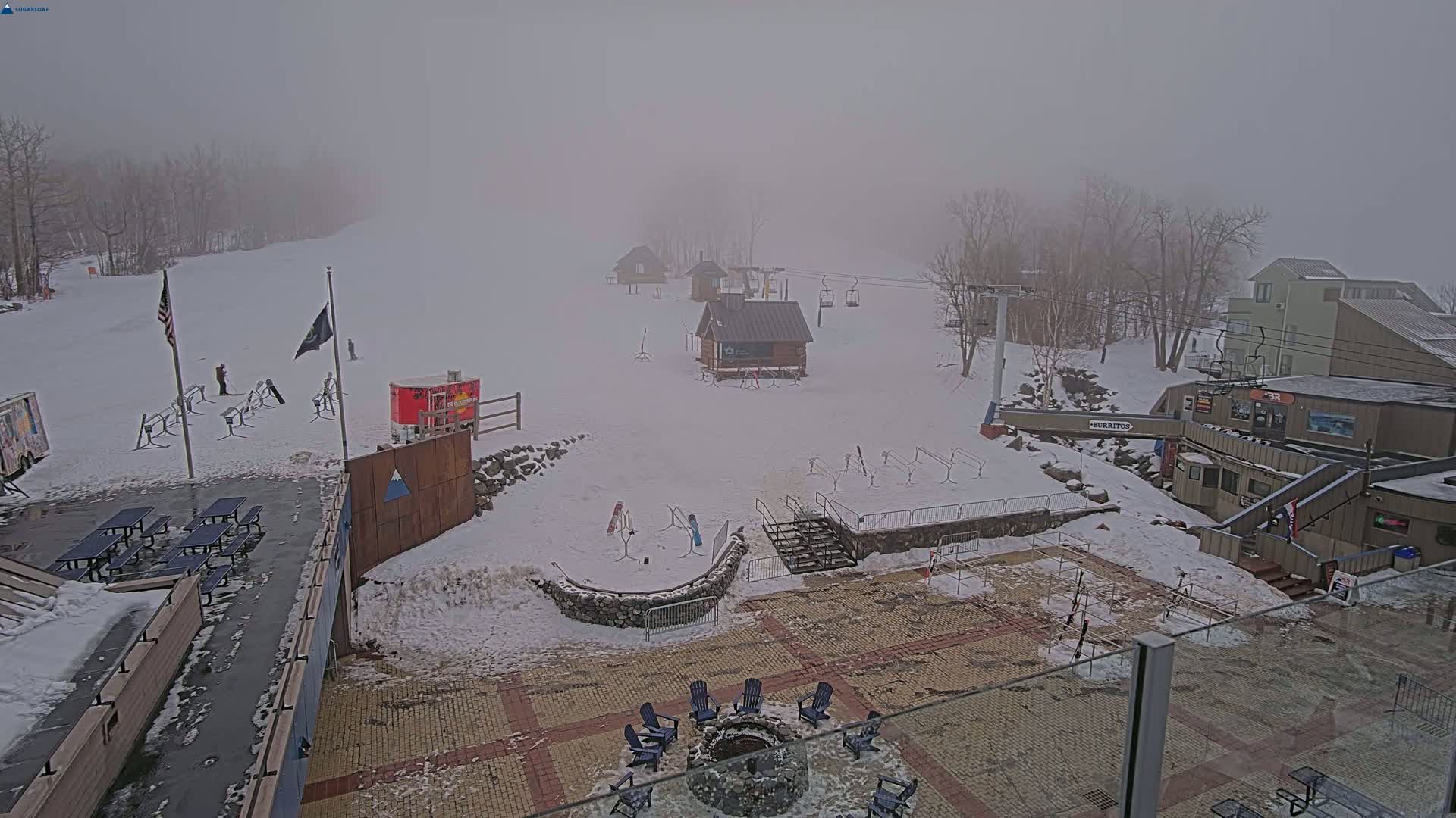 A snow-covered ski resort is shrouded in thick fog, with several small buildings, a chairlift, and outdoor seating areas visible.
