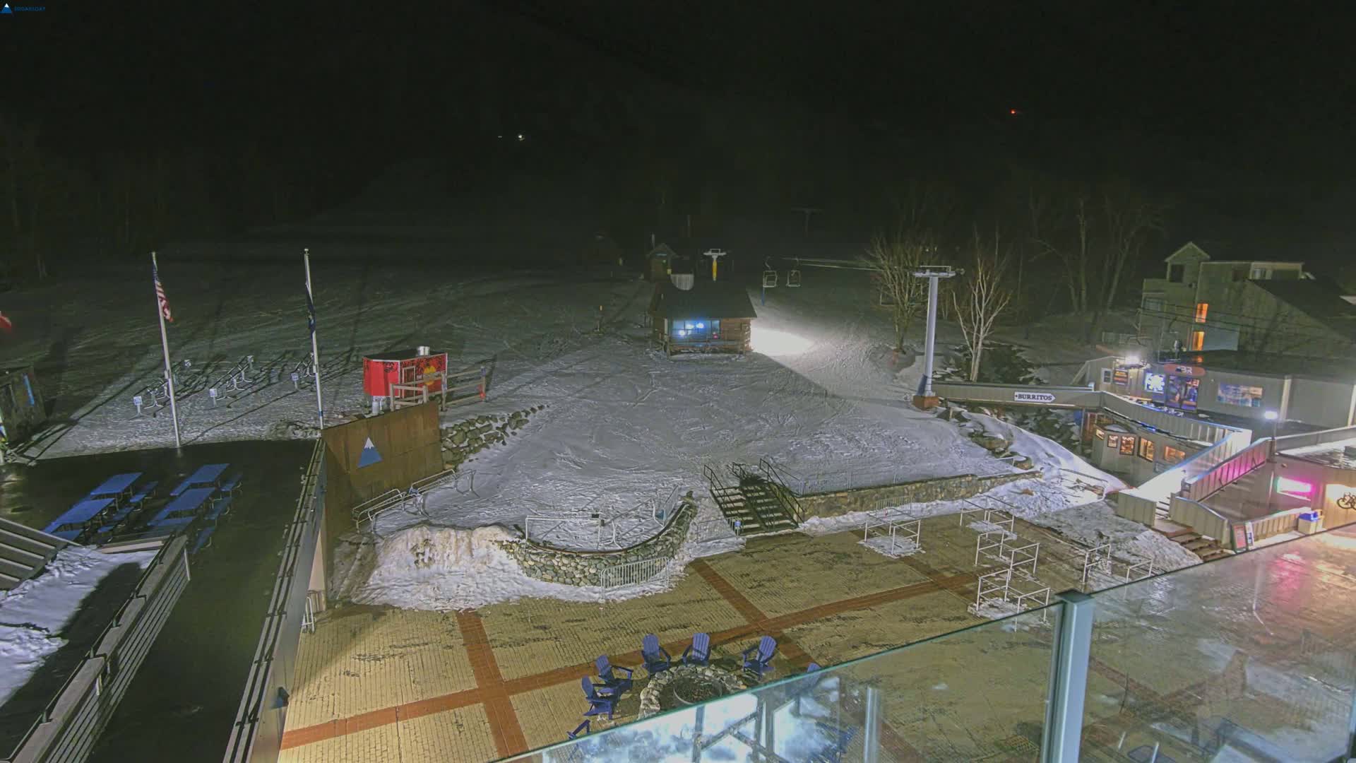 A snow-covered ski resort at night, illuminated by artificial lights, showing a ski lift, buildings, and outdoor seating areas.