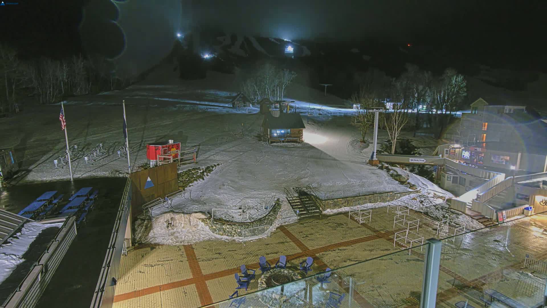 A snow-covered ski slope at night is illuminated by lights, with a patio area below featuring chairs around a fire pit and a building in the background.