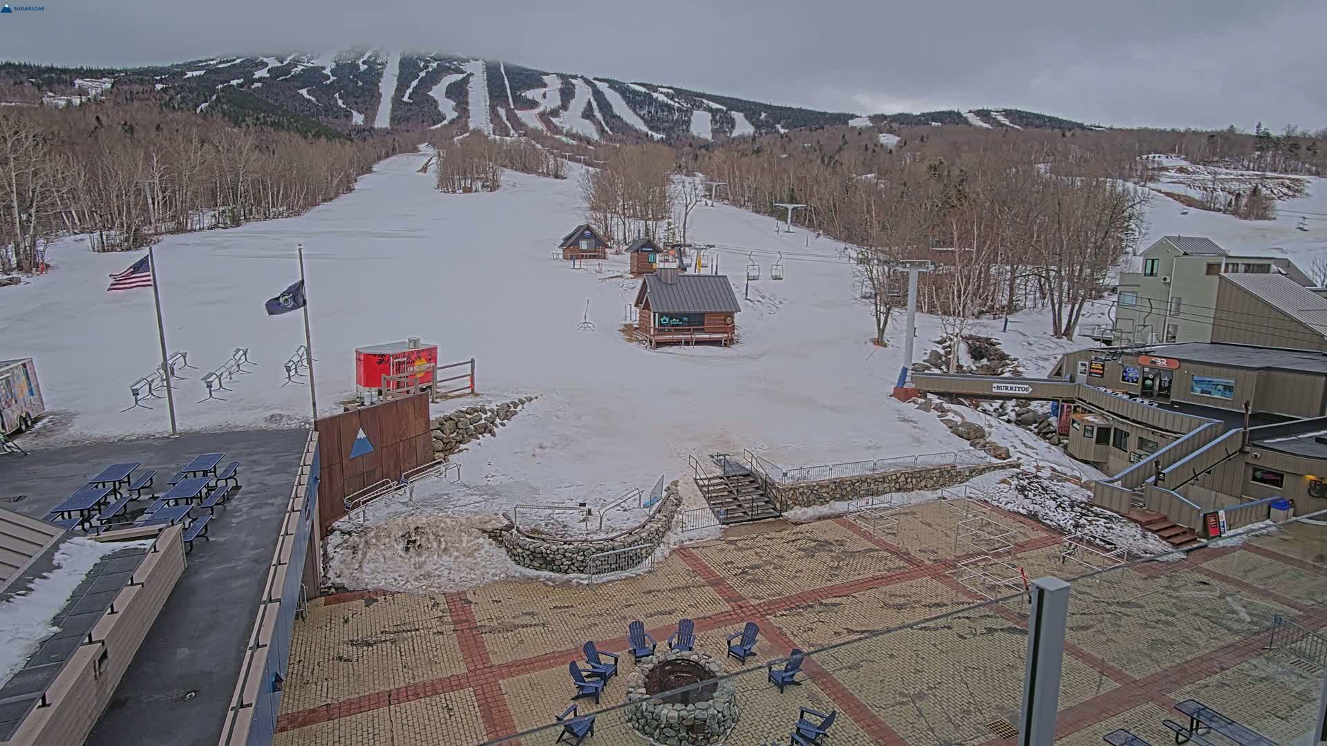 A snow-covered ski resort base area with several buildings, a fire pit, picnic tables, and a mountain with ski runs in the background under an overcast sky.