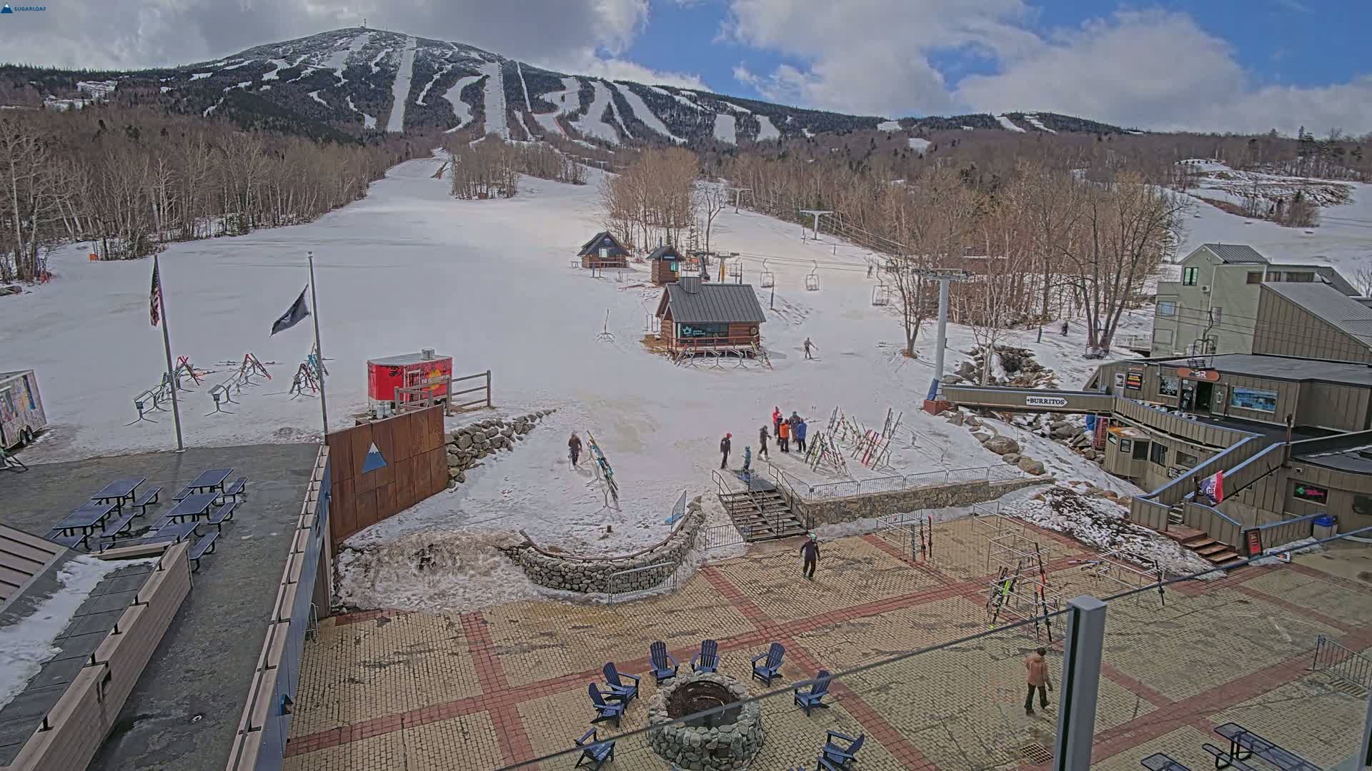 A mostly sunny day at a snow-covered ski resort shows a base area with several small buildings, a fire pit, picnic tables, and ski racks, all set against a backdrop of a large, snow-dusted mountain with ski runs.