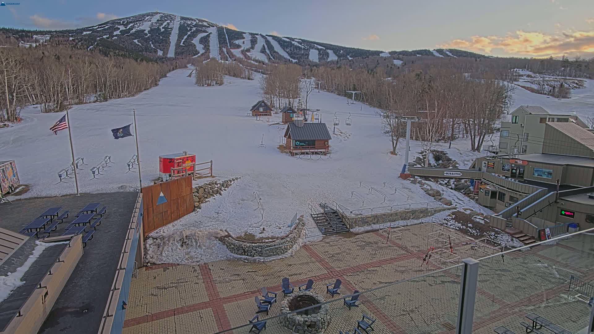 A snow-covered ski resort base area with several small buildings, picnic tables, a fire pit with chairs, and a mountain with ski runs in the background under a mostly clear, twilight sky.