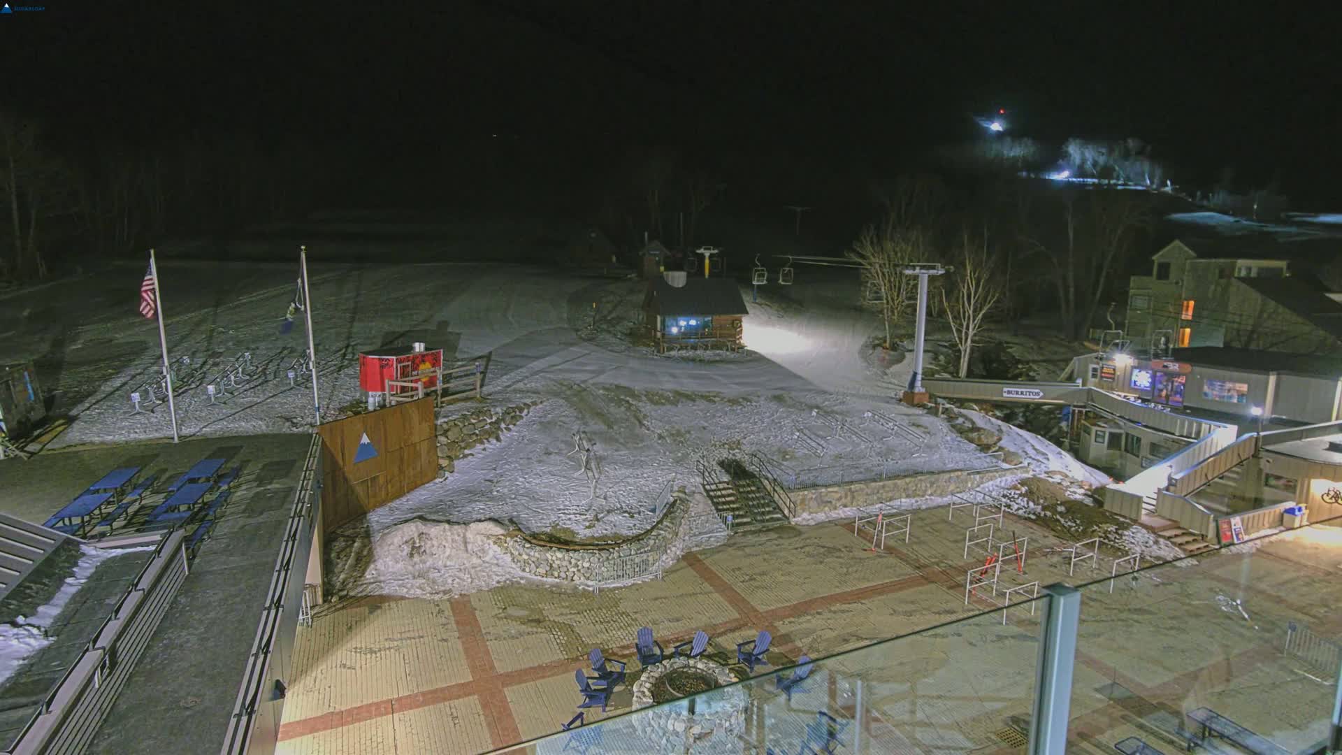A nighttime, snow-covered ski resort area is illuminated, showing a ski lift, a small lodge, outdoor seating, and a few buildings.