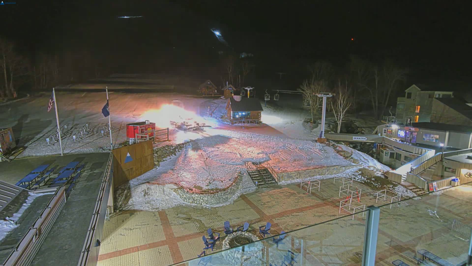 A nighttime view of a snow-covered ski resort base area with illuminated buildings, a ski lift, and fireworks going off.
