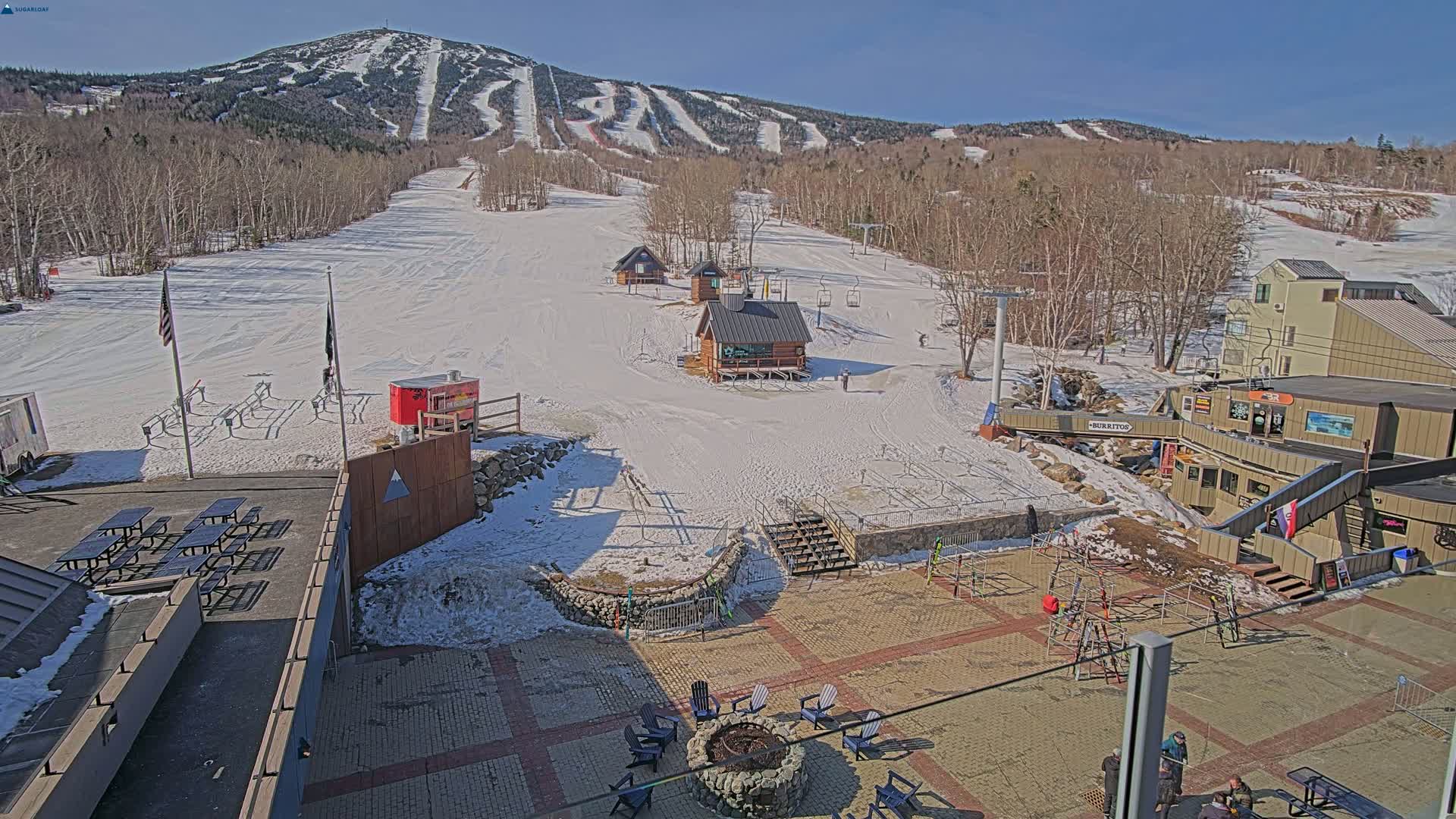 A snow-covered ski resort with several small buildings, picnic tables, a fire pit, and ski lifts, on a sunny day, with a mountain in the background.
