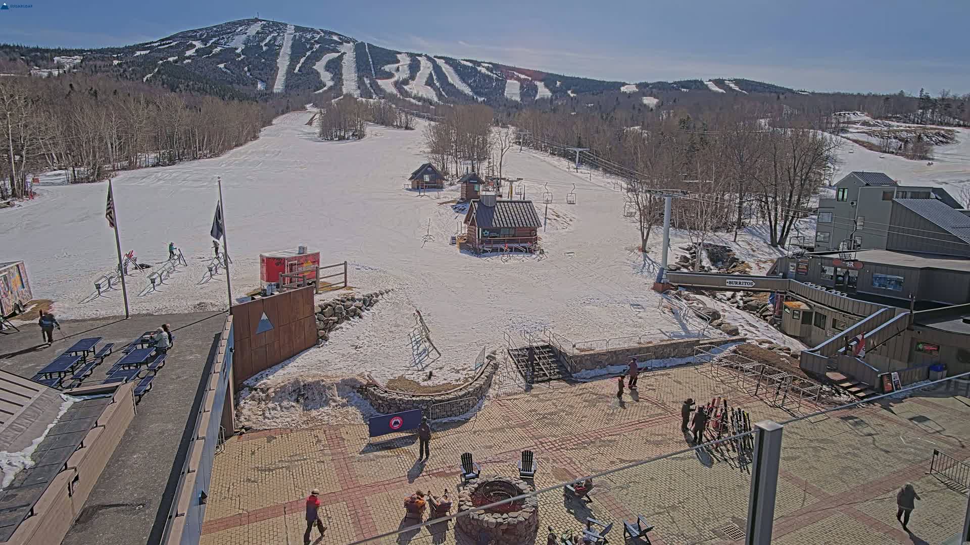A sunny day at a ski resort shows snow-covered slopes, ski lifts, small buildings, and people enjoying the outdoor patio area.