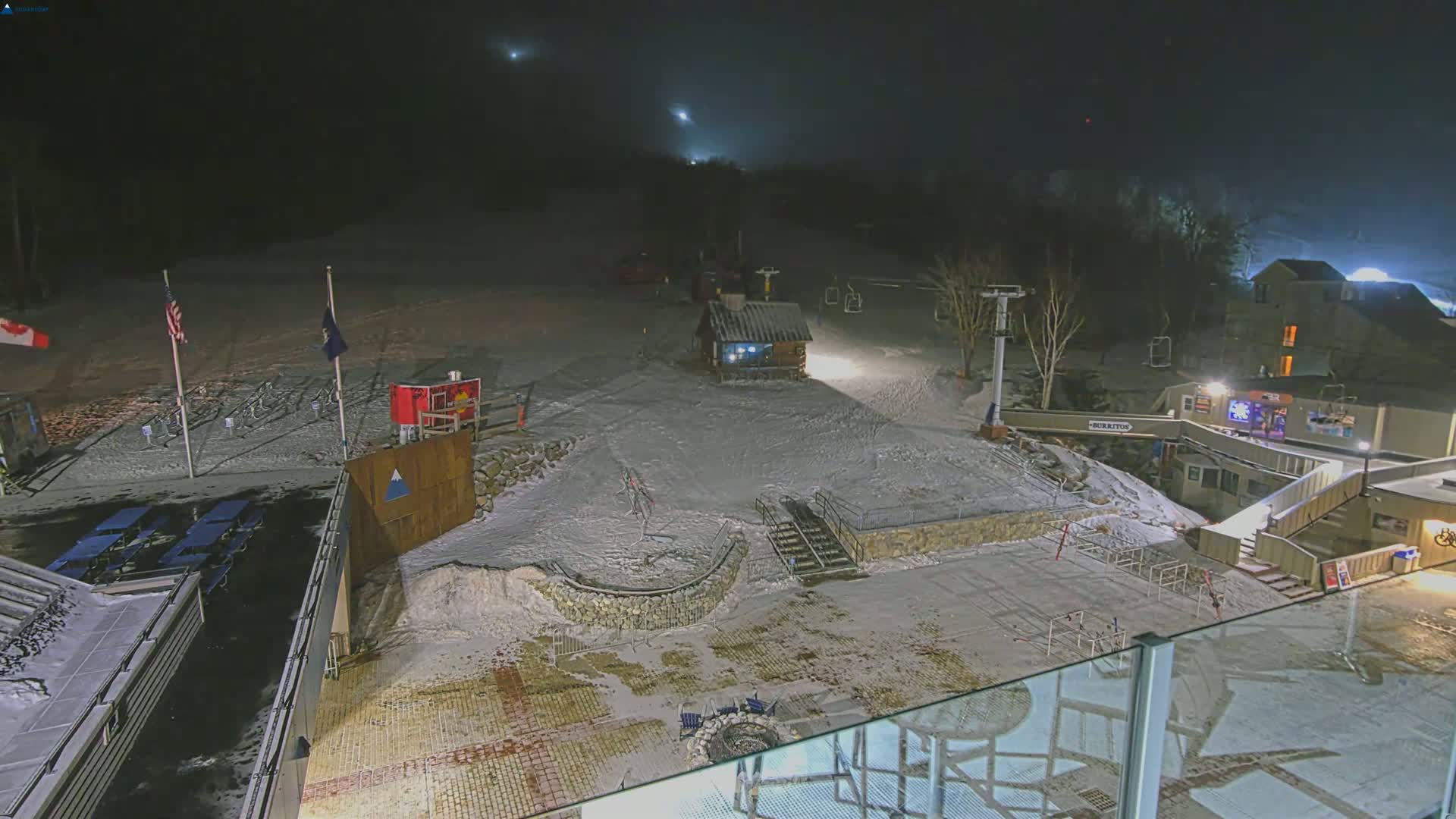 A snow-covered ski resort at night, illuminated by artificial lights, with a small building and ski lift visible in the hazy atmosphere.