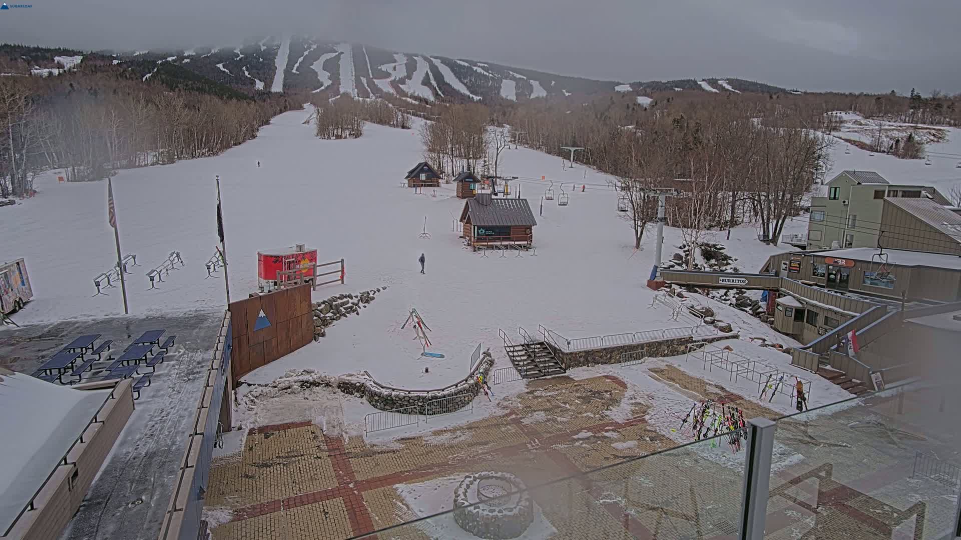 A snow-covered ski resort with several buildings, ski lifts, and picnic tables, under an overcast sky.