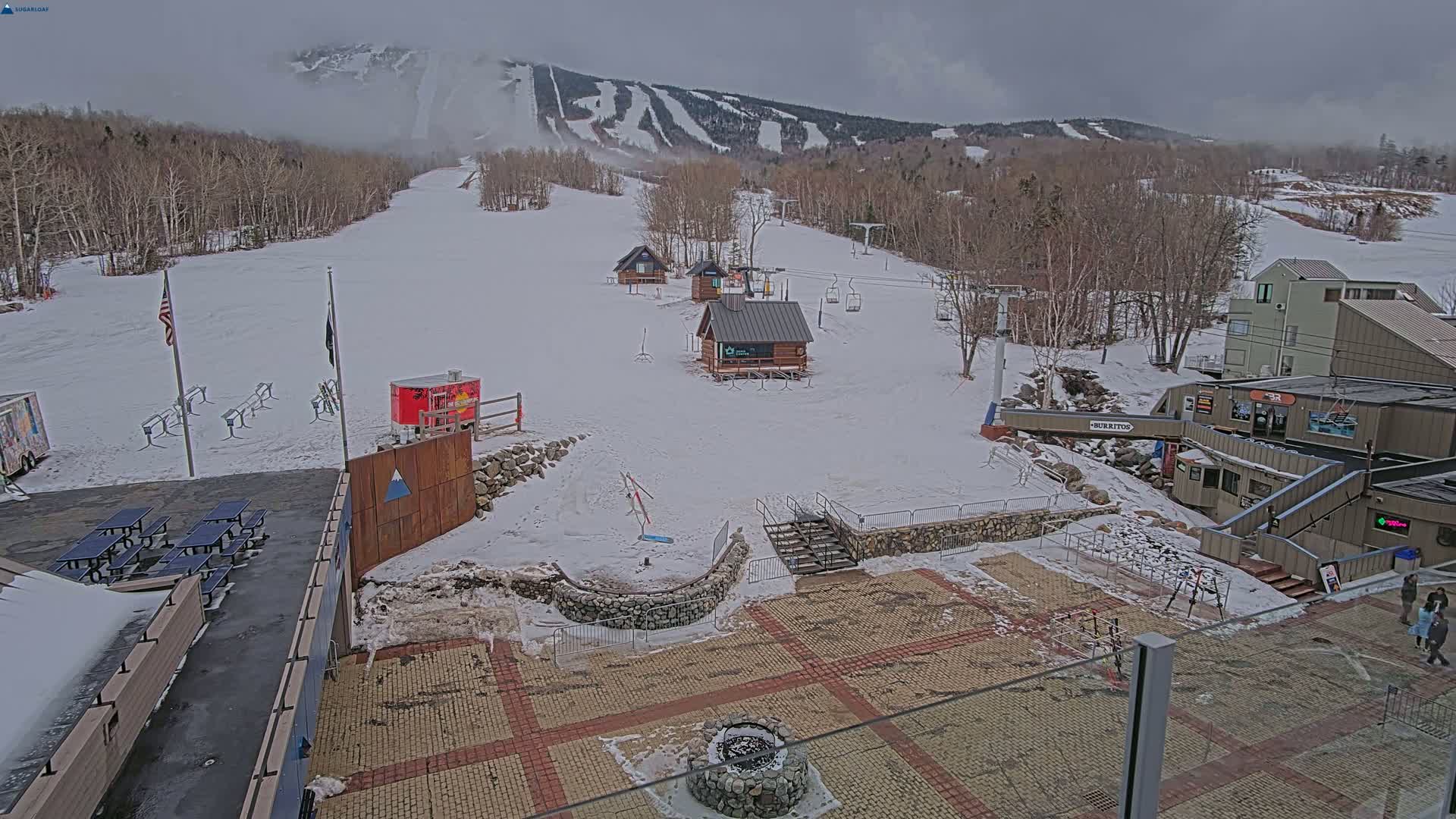 A snow-covered ski resort base area with several small buildings, picnic tables, and ski lifts, under an overcast sky.