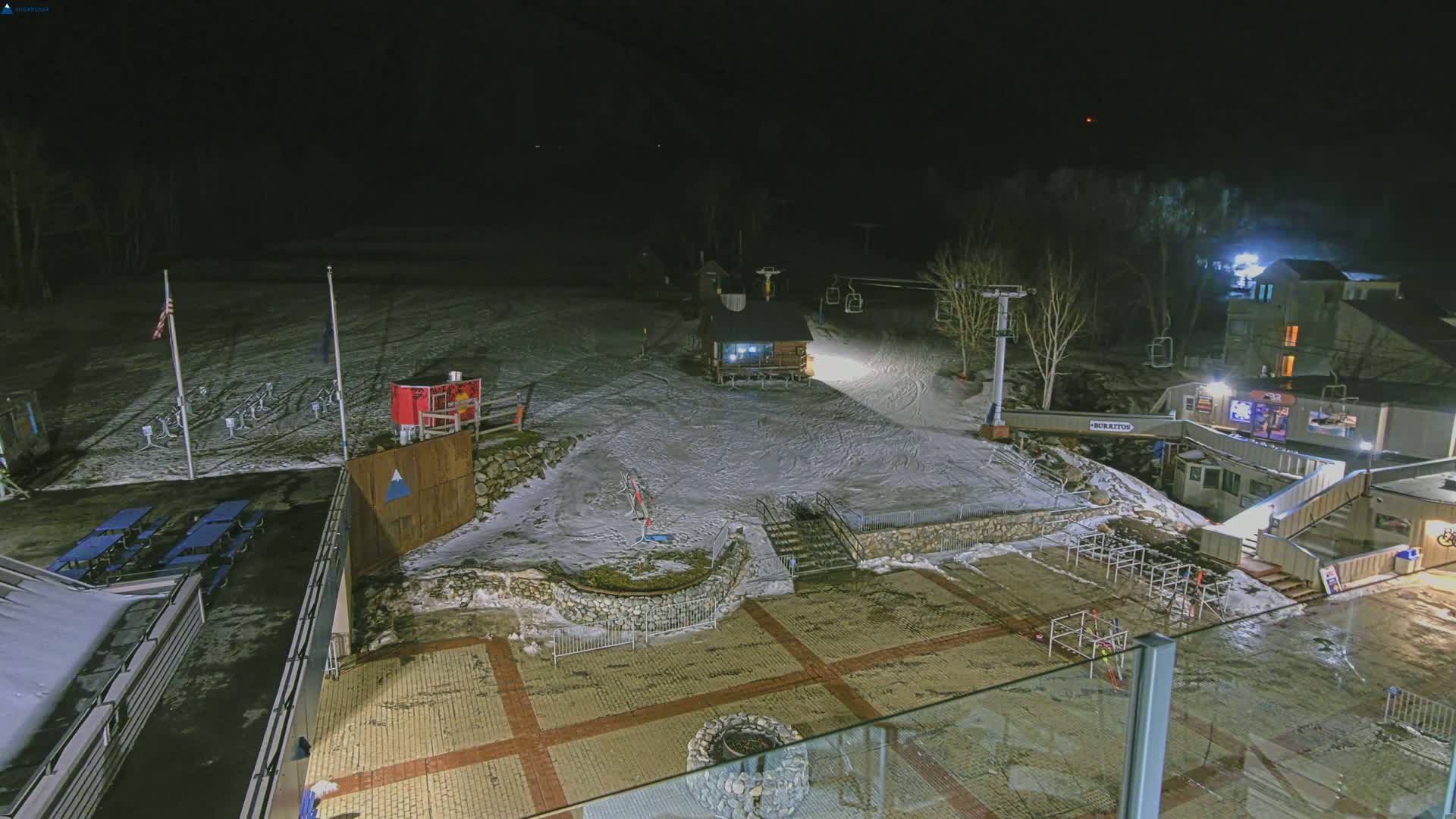 A nighttime, snow-covered ski resort scene shows a ski lift, a small building, and a paved area with some picnic tables.