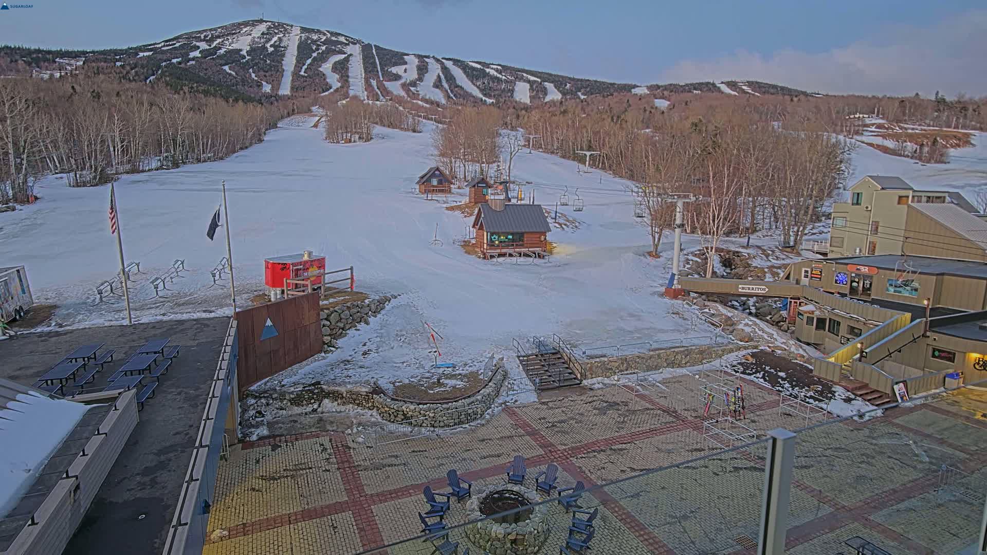 A snow-covered ski resort base area with several buildings, a fire pit, picnic tables, and ski lifts against a backdrop of a snow-covered mountain under a twilight sky.