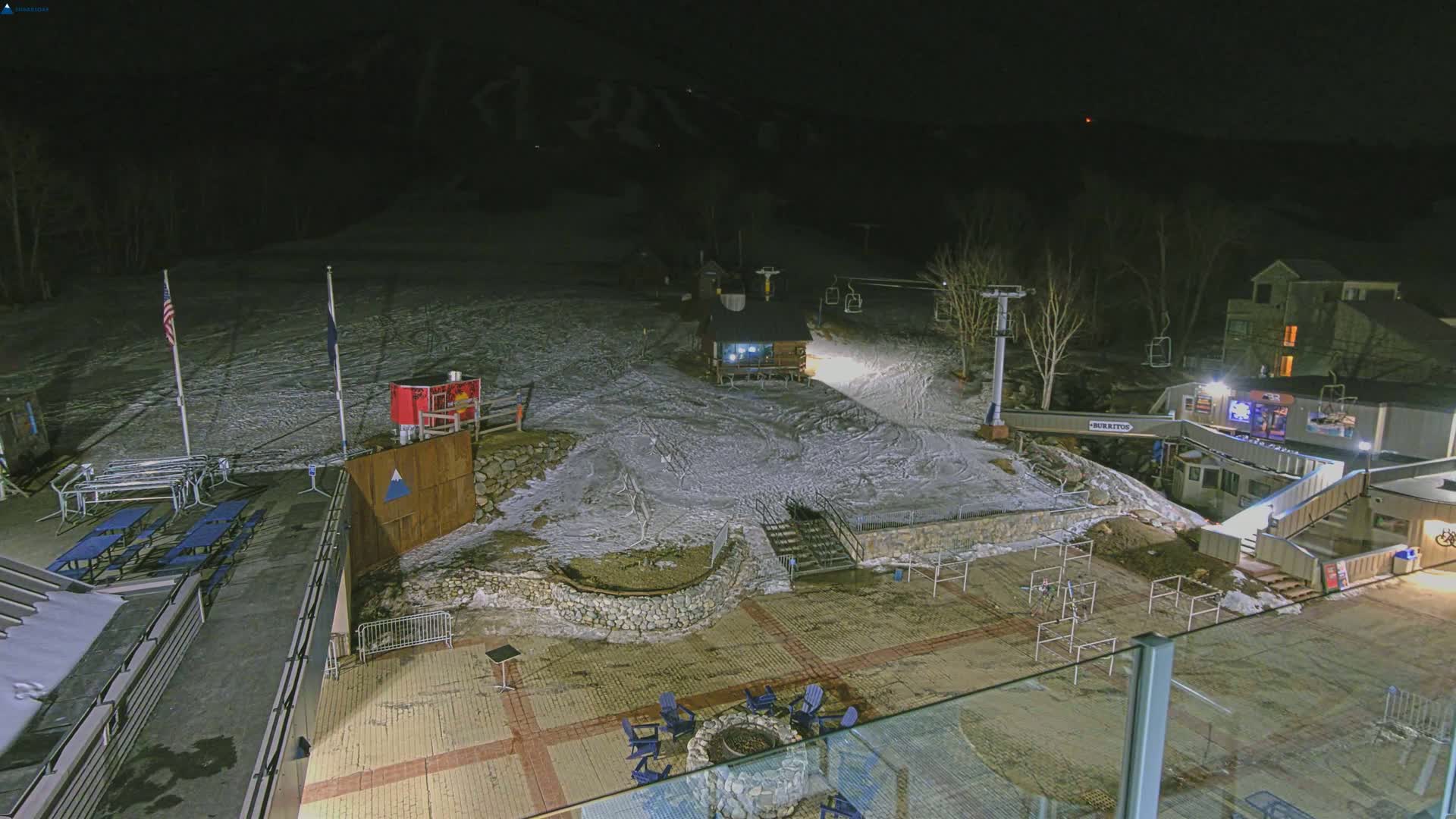 A nighttime view of a snow-covered ski resort base area with a chairlift, buildings, picnic tables, and a fire pit.