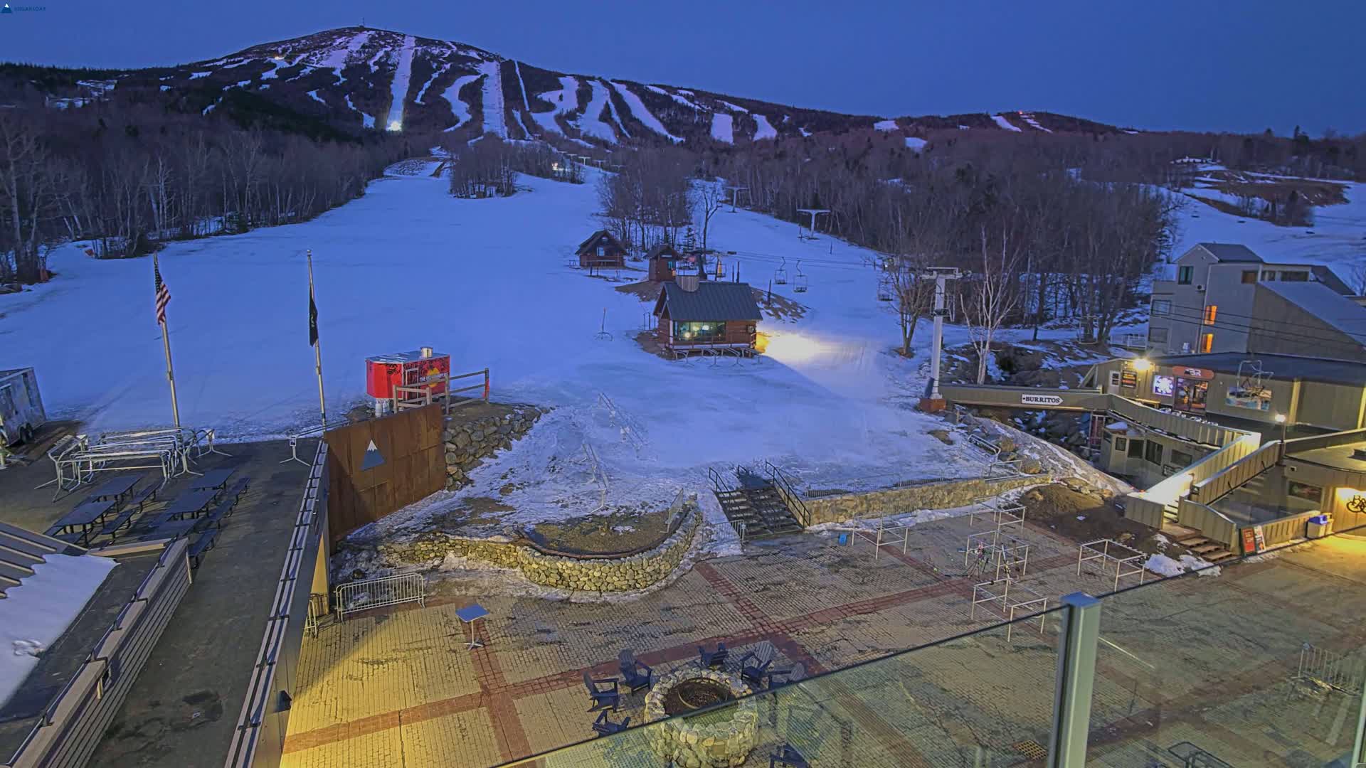 A snow-covered ski resort at twilight, with several small buildings, picnic tables, and a fire pit visible in the foreground, and a mountain with ski slopes in the background.