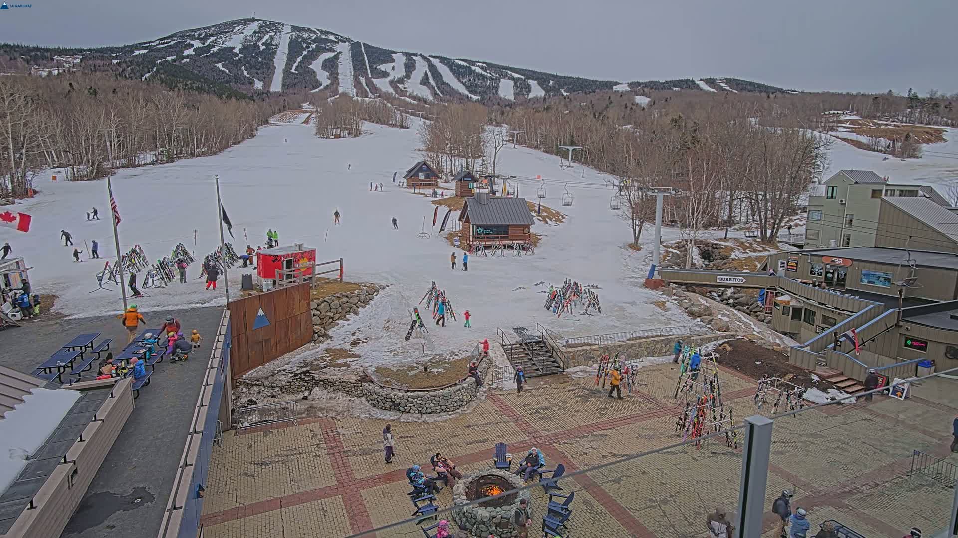 A snow-covered ski resort base area with people gathered around a fire pit, skiers on the slopes, and ski equipment stacked in various locations, under a cloudy sky.