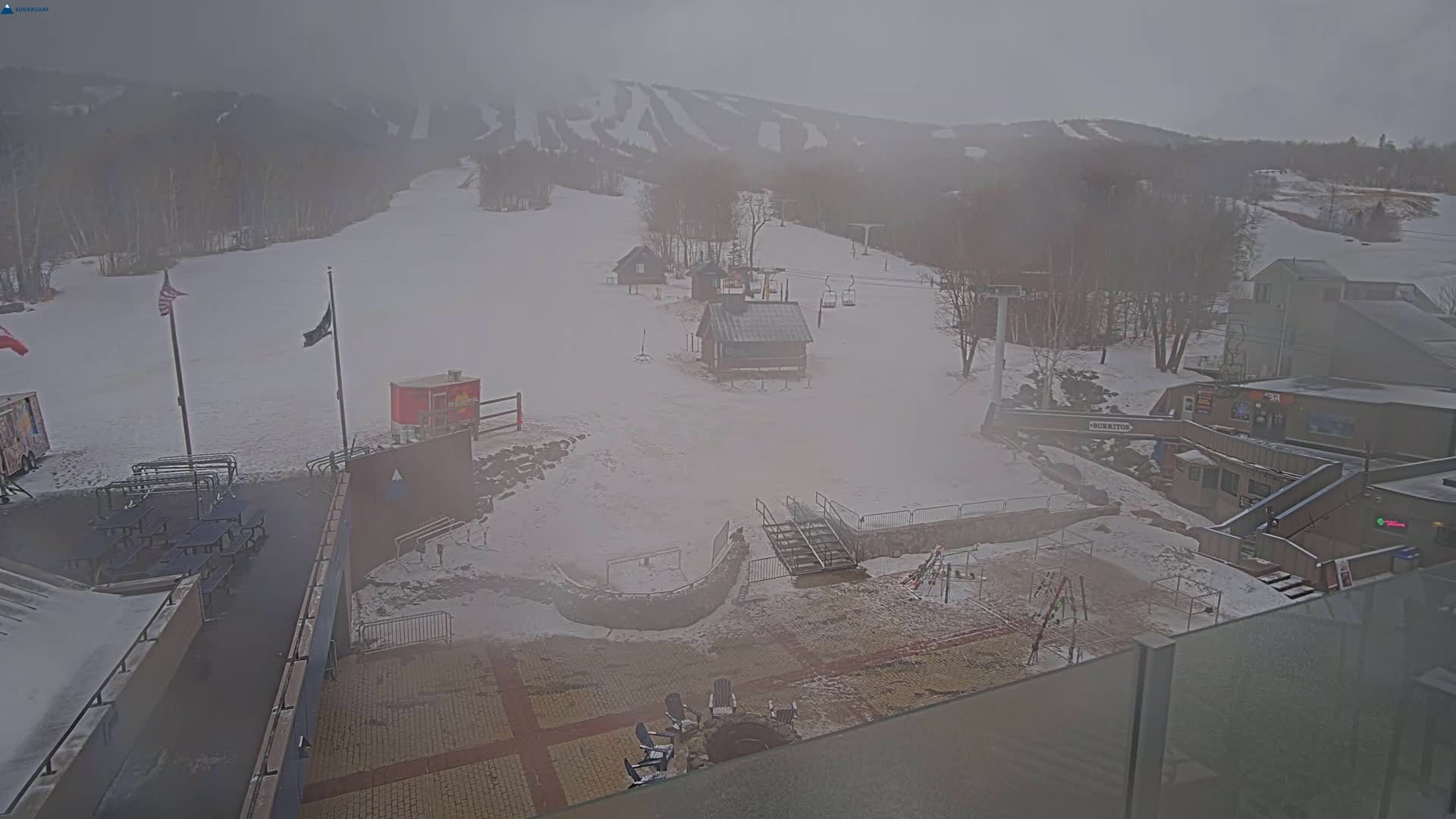 A foggy view of a snow-covered ski resort base area with several buildings, ski lifts, and outdoor seating areas.