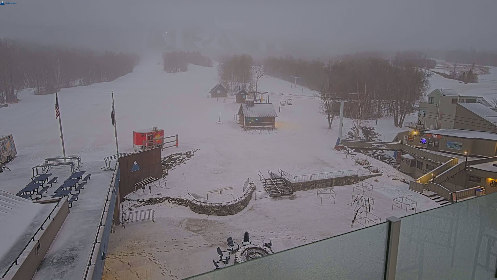 A snowy ski resort is shrouded in fog, featuring several buildings, a chairlift, picnic tables, and a fire pit.
