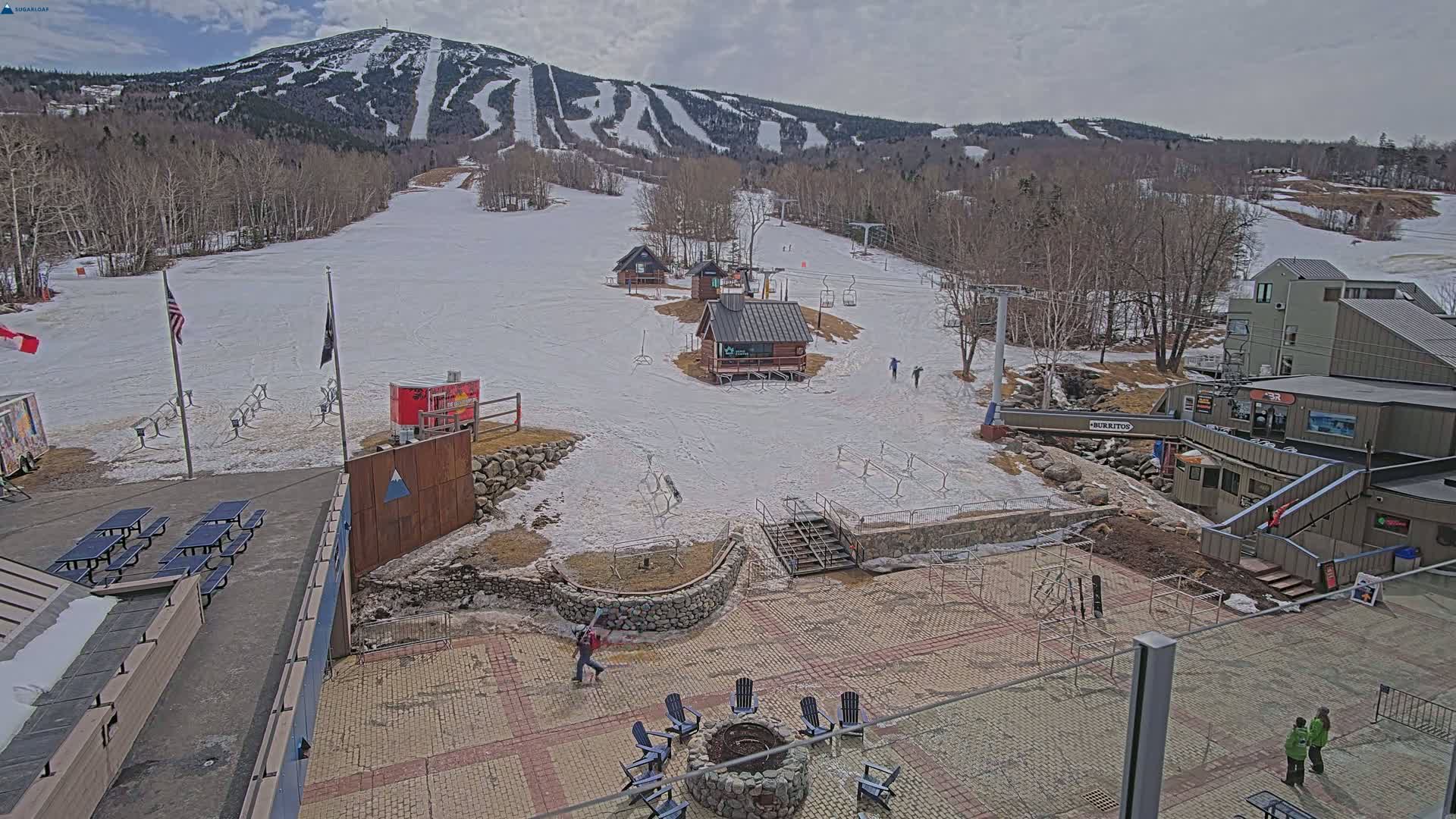 A snow-covered ski resort base area with several buildings, picnic tables, a fire pit, and a mountain with ski runs in the background under a cloudy sky.