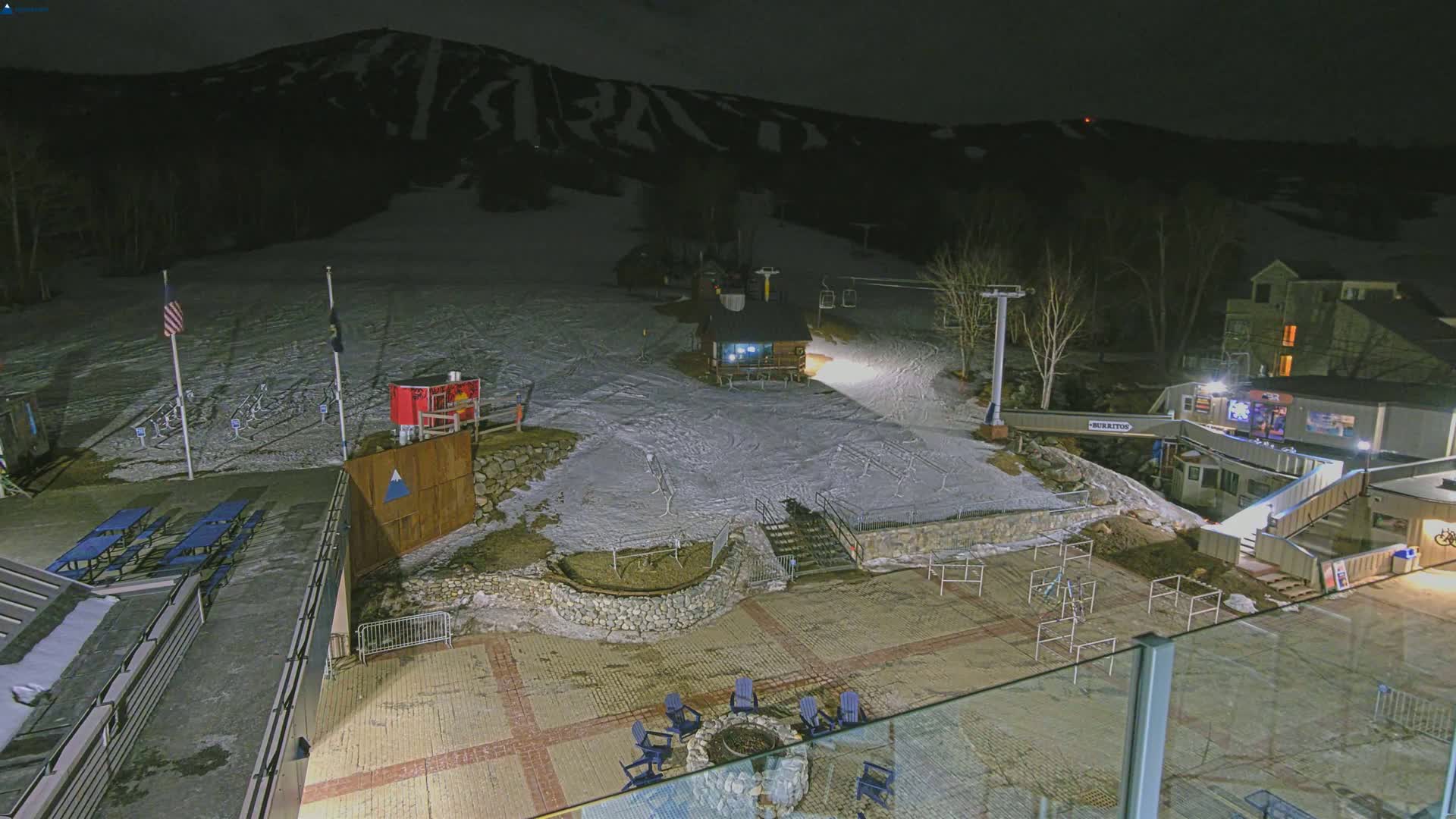 A nighttime view of a snow-covered ski resort base area with buildings, a ski lift, and outdoor seating around a fire pit under clear skies.