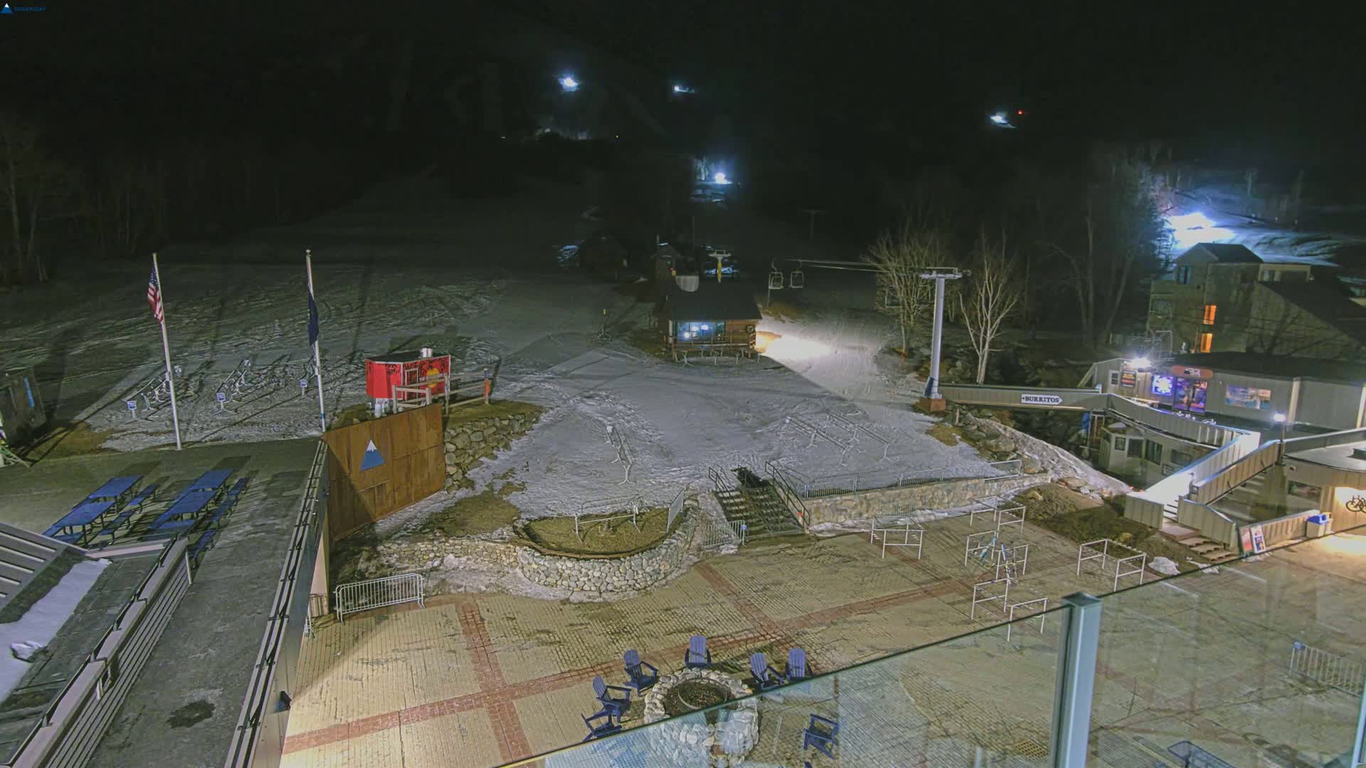 A snow-covered ski slope at night, illuminated by artificial lights, with a base area featuring picnic tables, a fire pit, and buildings.