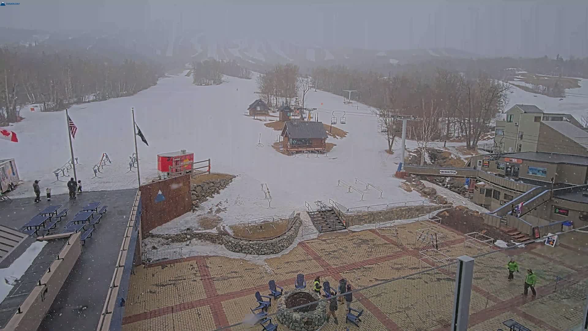 A snow-covered ski resort base area with several buildings, a fire pit, and picnic tables, under snowy conditions.