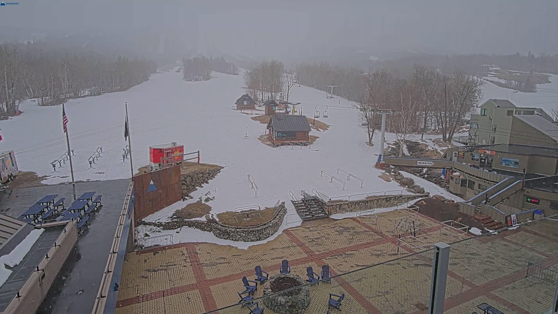 A snow-covered ski slope with several small buildings, a chairlift, and a paved patio area with picnic tables and a fire pit, all under a foggy, overcast sky.
