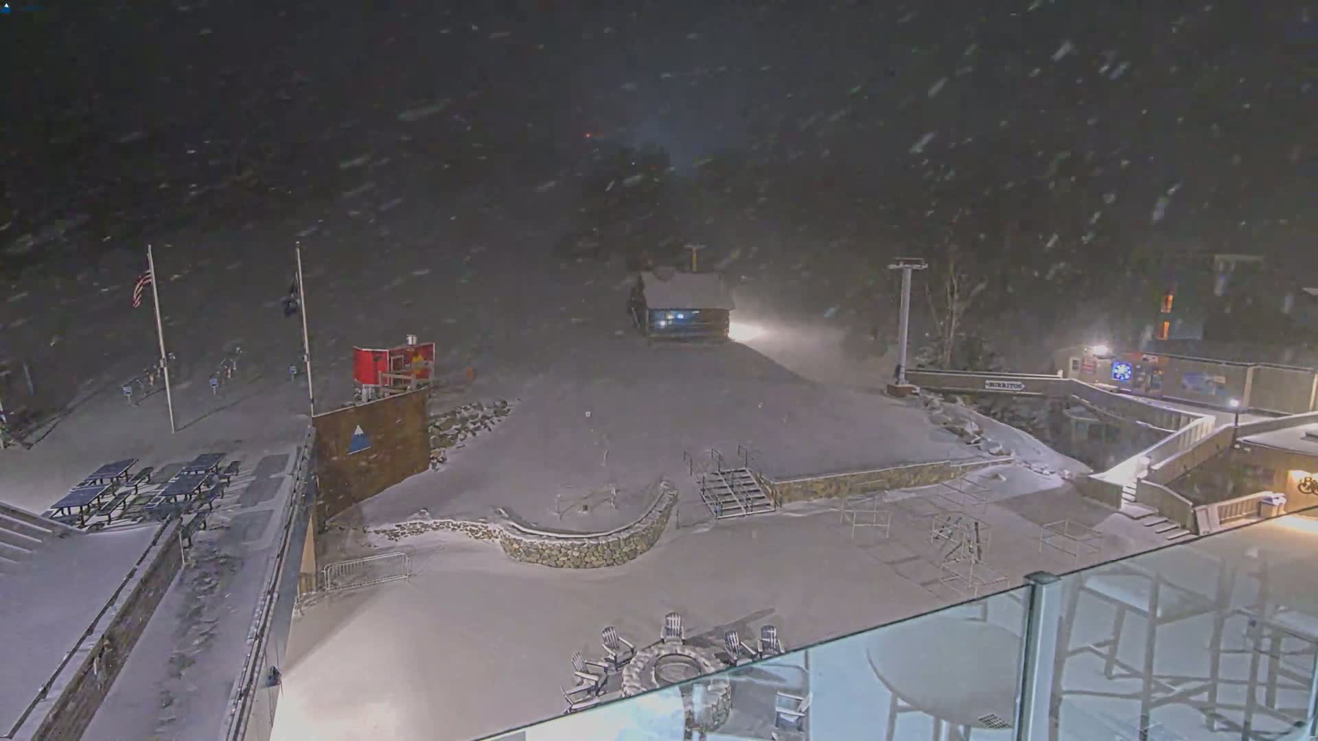 A snow-covered outdoor area at night, with falling snow, features picnic tables, a small building, and a fire pit with chairs around it.
