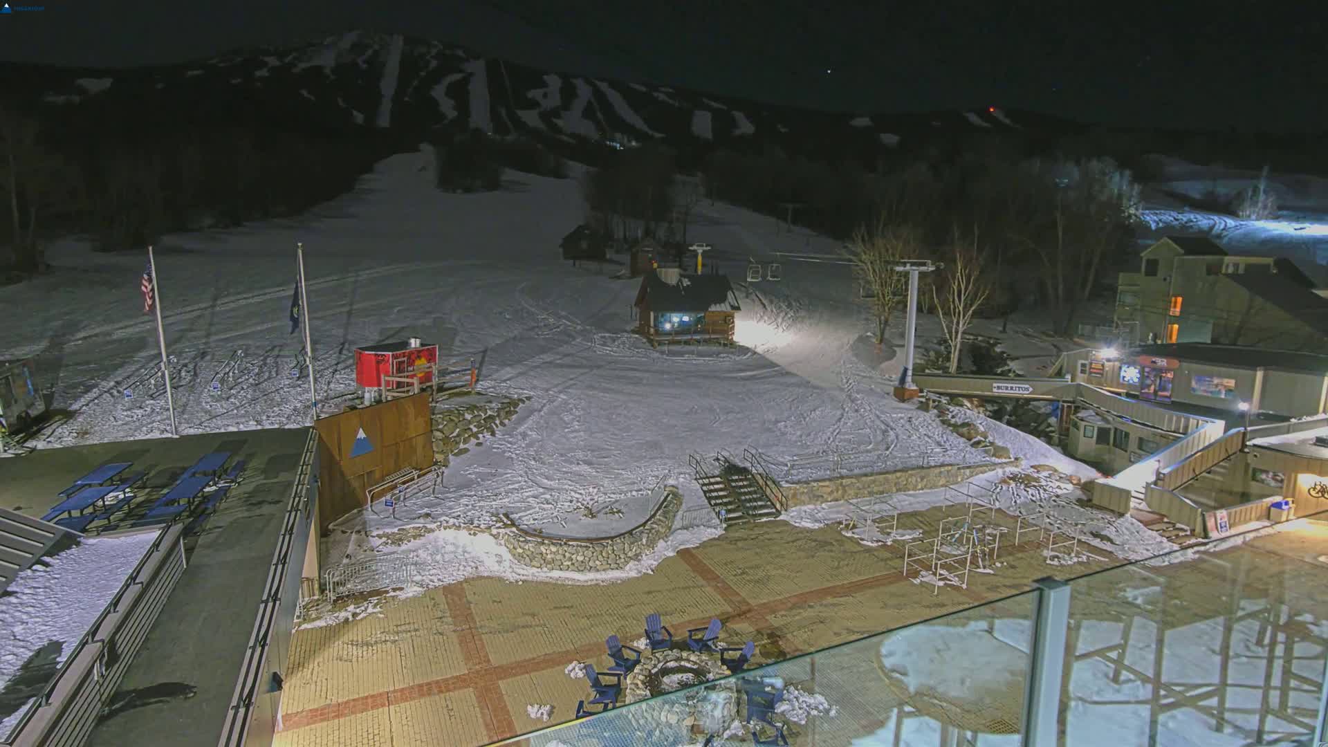 A nighttime view of a snow-covered ski resort base area with a fire pit and chairs, a ski lift, and buildings under a clear, dark sky.