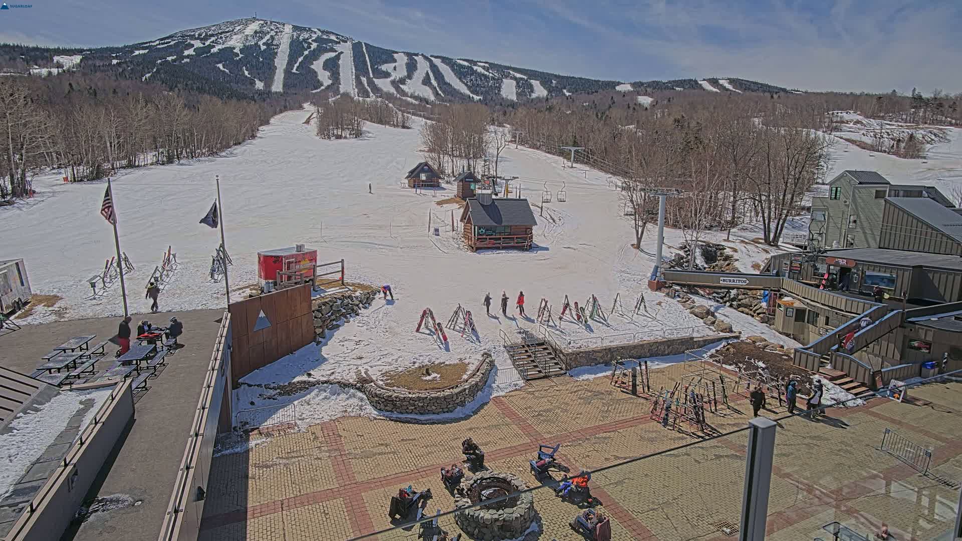 A sunny, snow-covered ski resort base area with several people gathered around a fire pit and others walking on paved paths, featuring ski lifts, small buildings, and a mountain in the background.