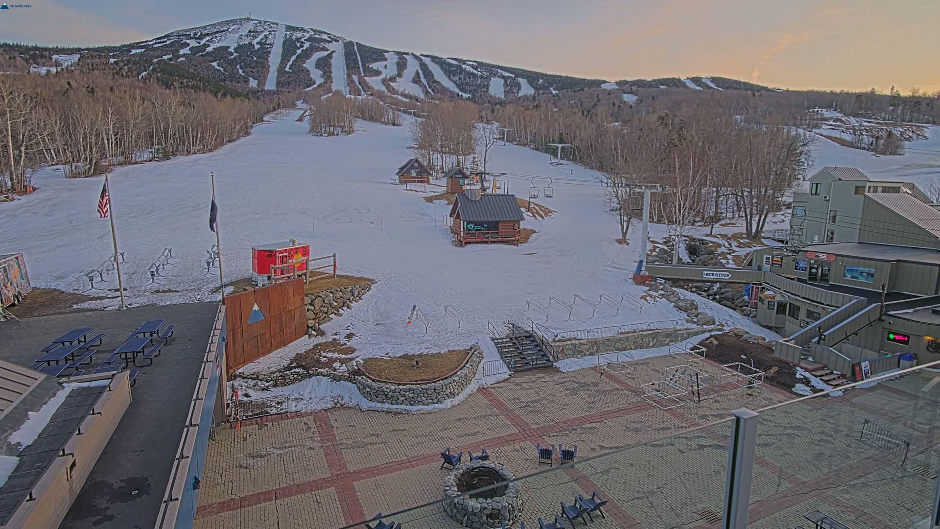 A snow-covered ski resort base area with several small buildings, picnic tables, and a fire pit, situated at the foot of a mountain with ski runs visible under a twilight sky.