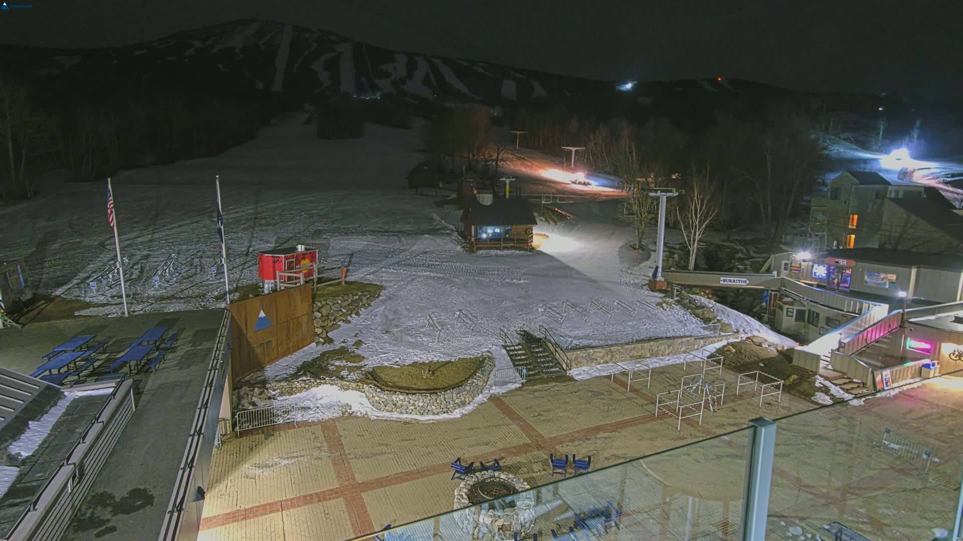 A snow-covered ski slope at night, illuminated by lights, with a small building and outdoor seating area below.