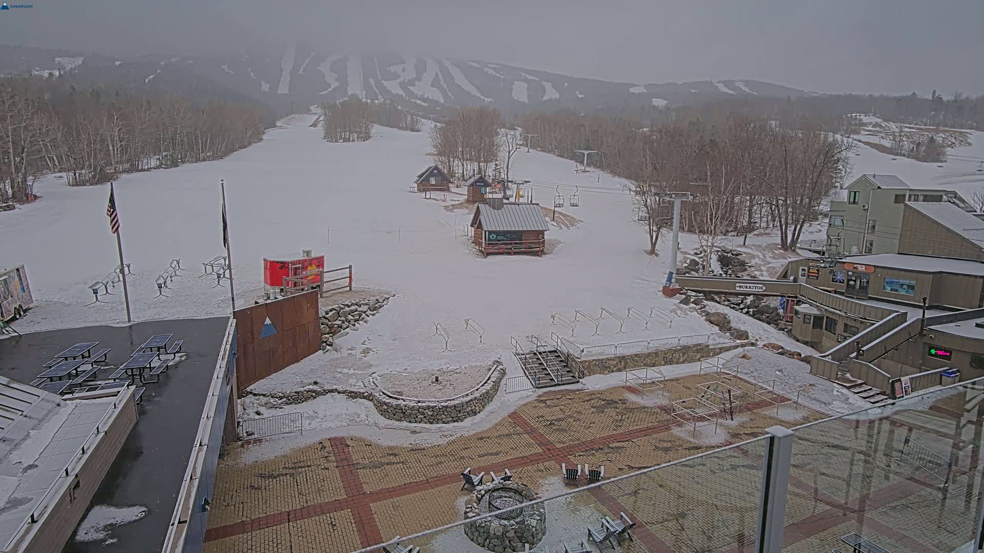 A snow-covered ski resort base area with several buildings, picnic tables, and a ski lift, all under an overcast sky.