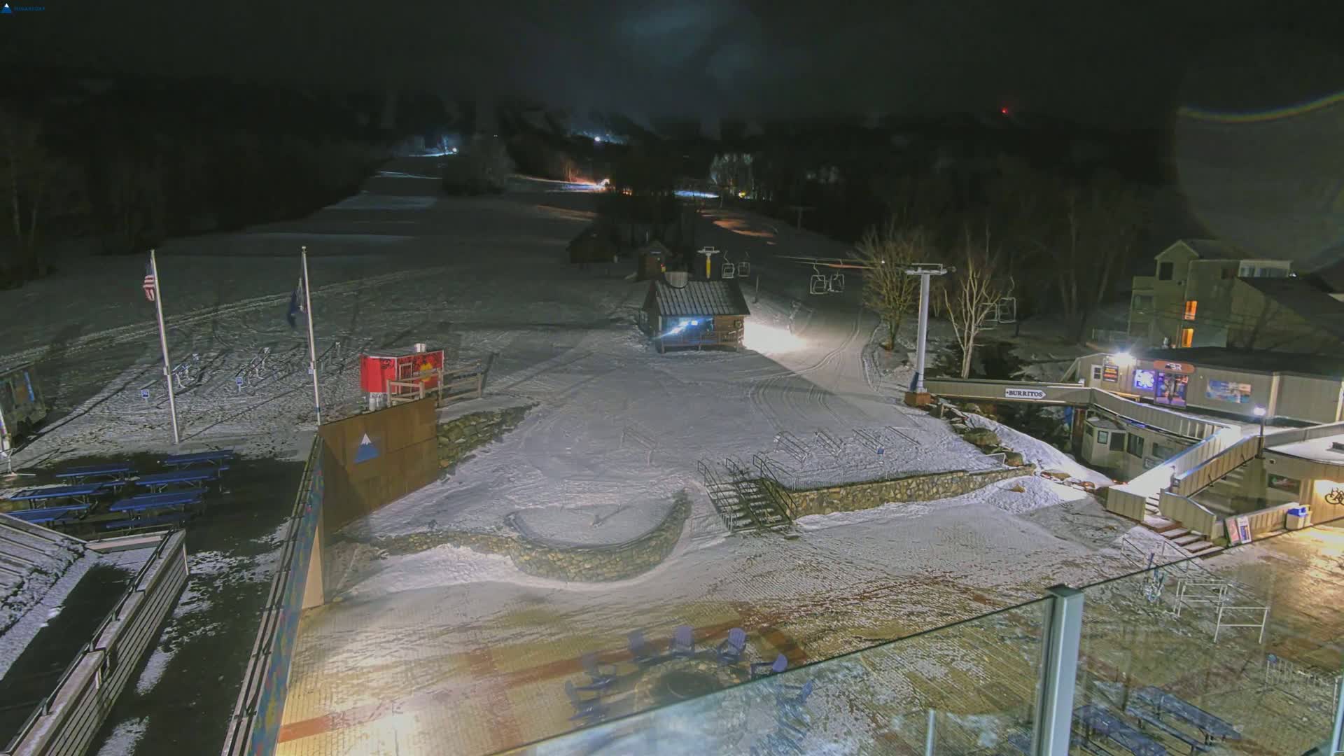 A snow-covered ski slope at night, illuminated by artificial lights, with a small building and ski lift visible.