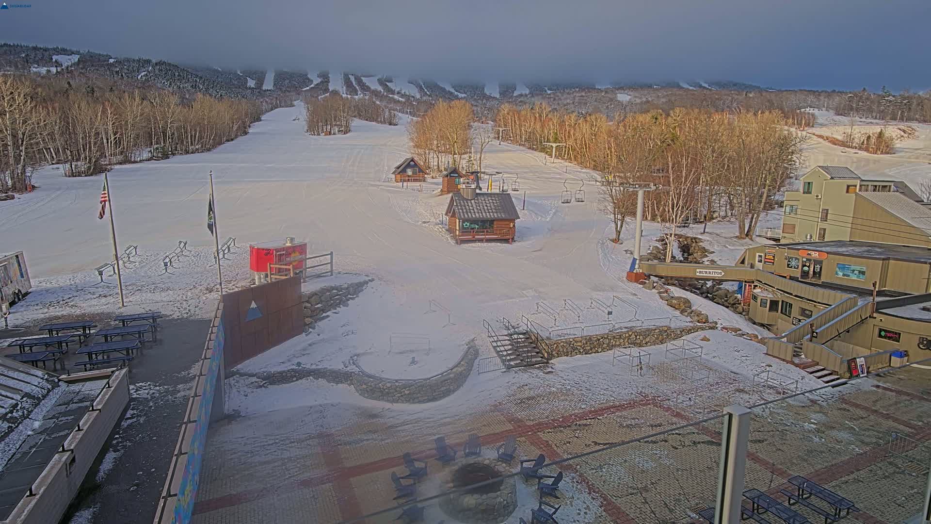 A snow-covered ski resort base area with several buildings, picnic tables, a fire pit, and ski lifts under a cloudy sky.