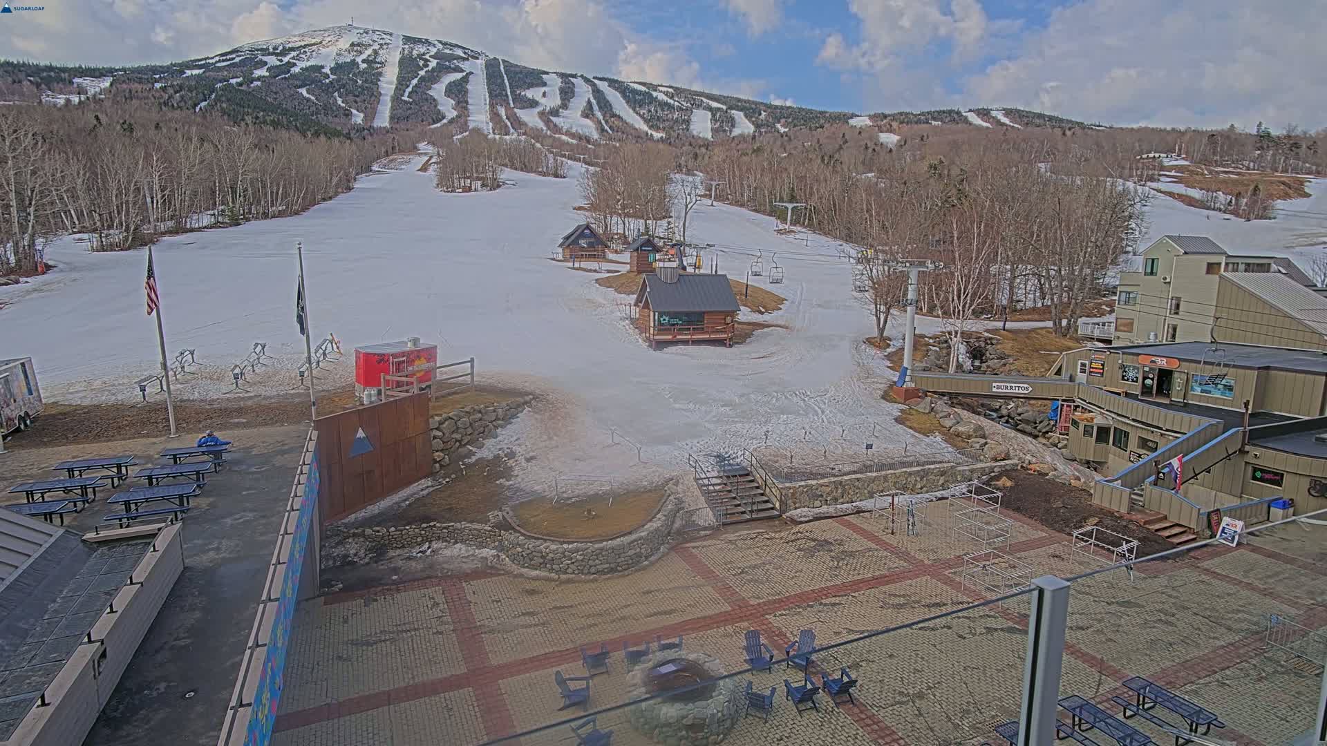 A snow-covered ski resort base area with several buildings, picnic tables, and a fire pit, situated beneath a partly cloudy sky and a mountain with ski trails.