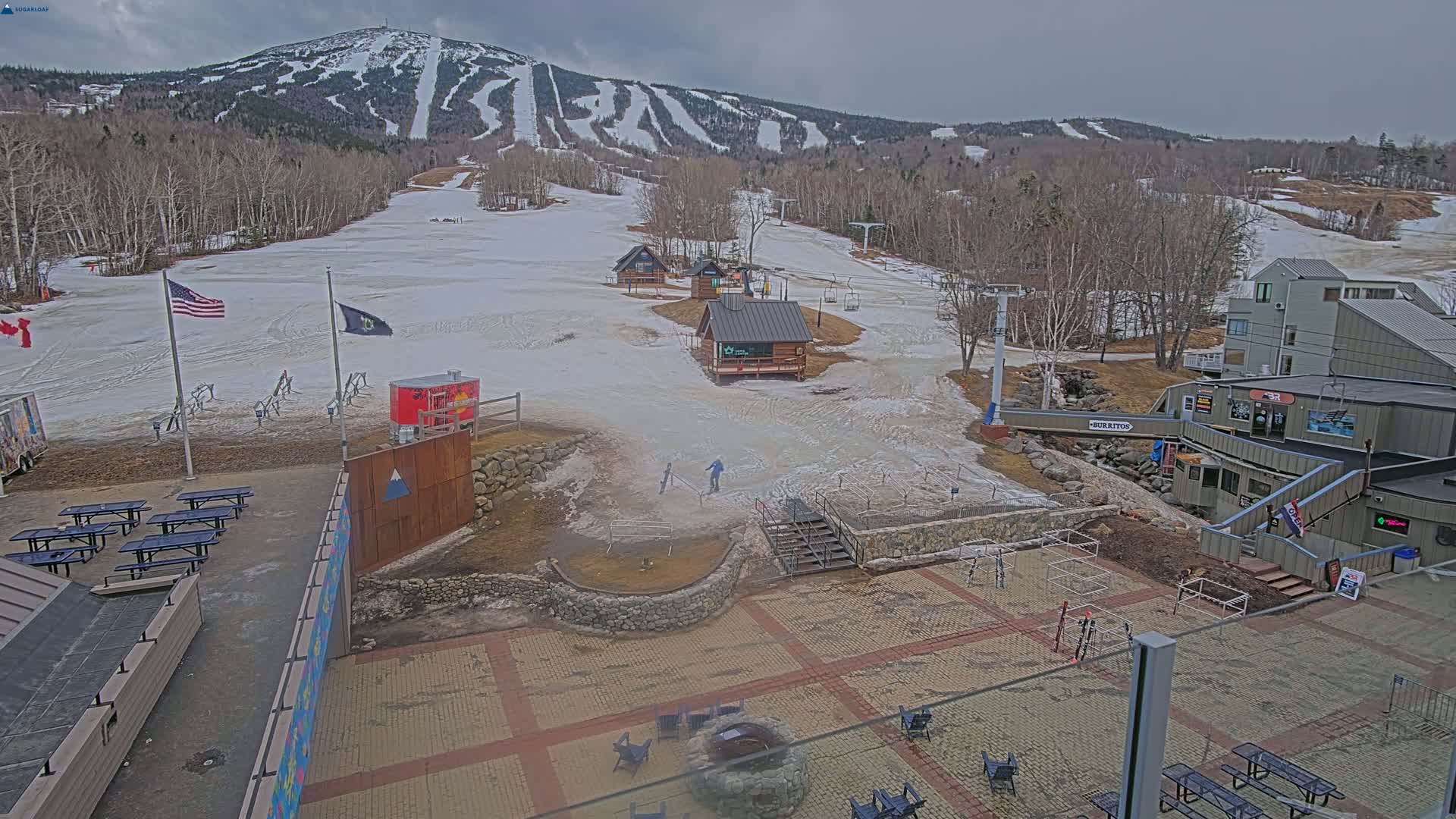 A snow-covered ski resort with several buildings, picnic tables, and a fire pit, under an overcast sky.
