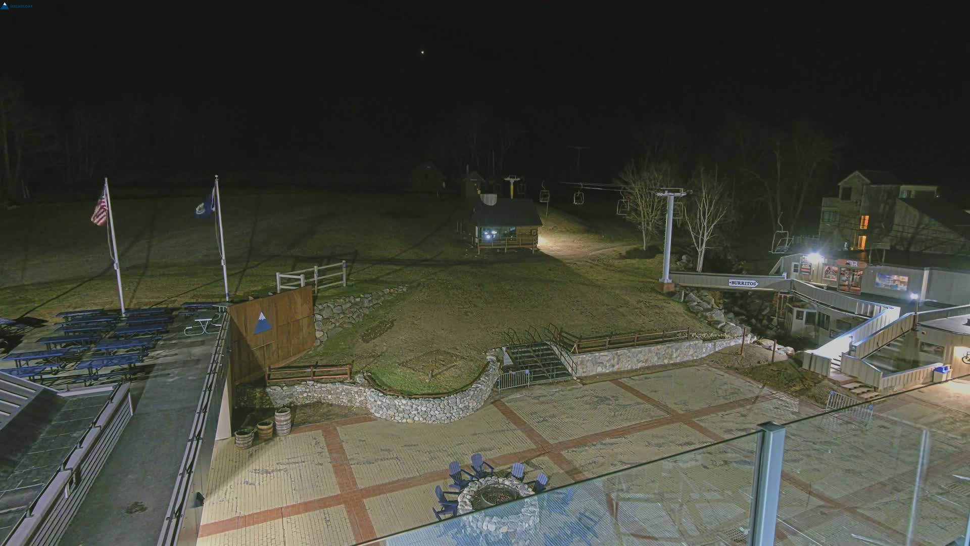 An outdoor nighttime scene shows a paved area with picnic tables, a fire pit, a ski lift, and a building, all under a dark sky.