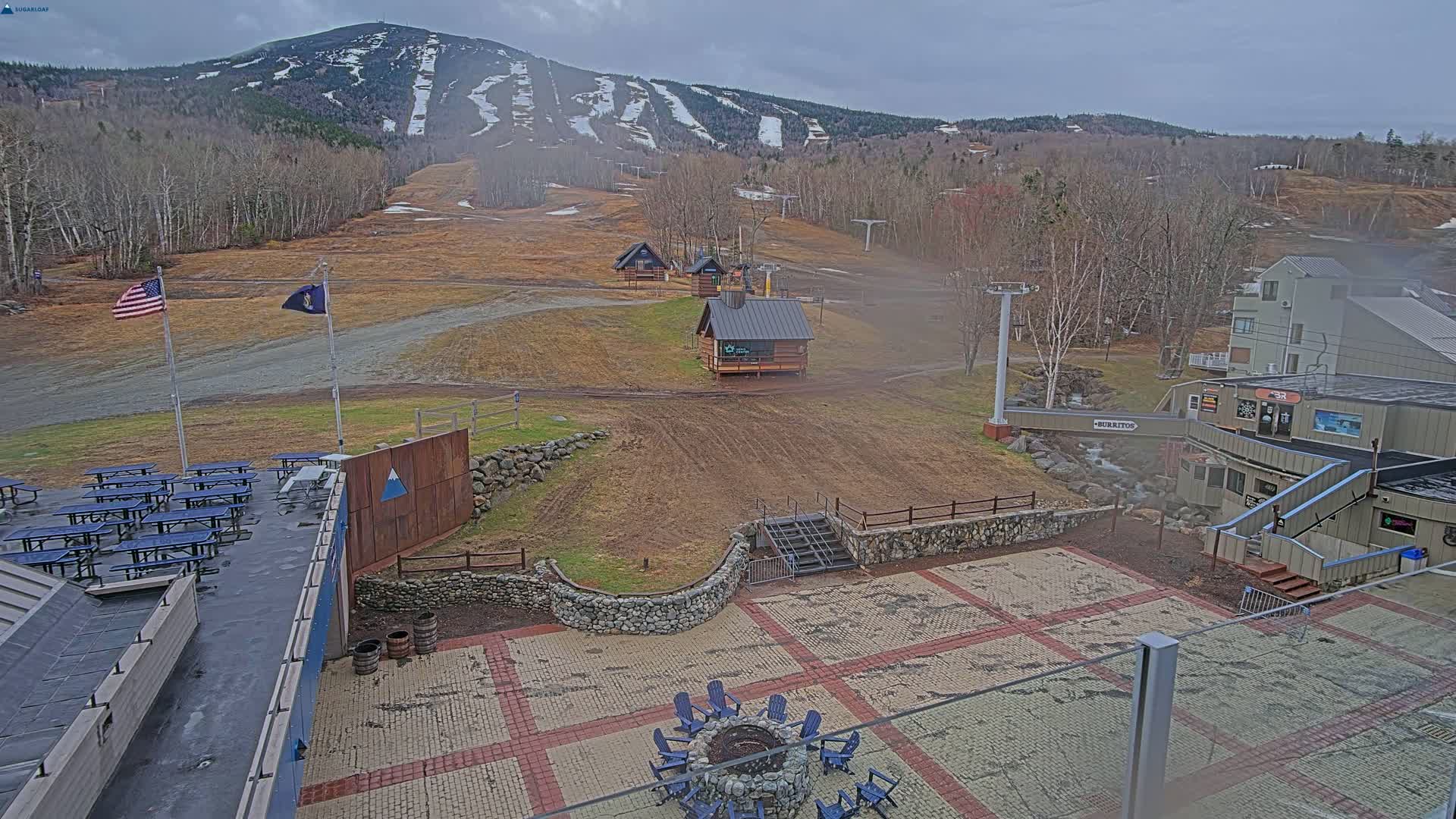 A paved patio area with picnic tables and a fire pit sits in front of a ski resort building, overlooking brown ski slopes and a snow-dusted mountain under an overcast sky.