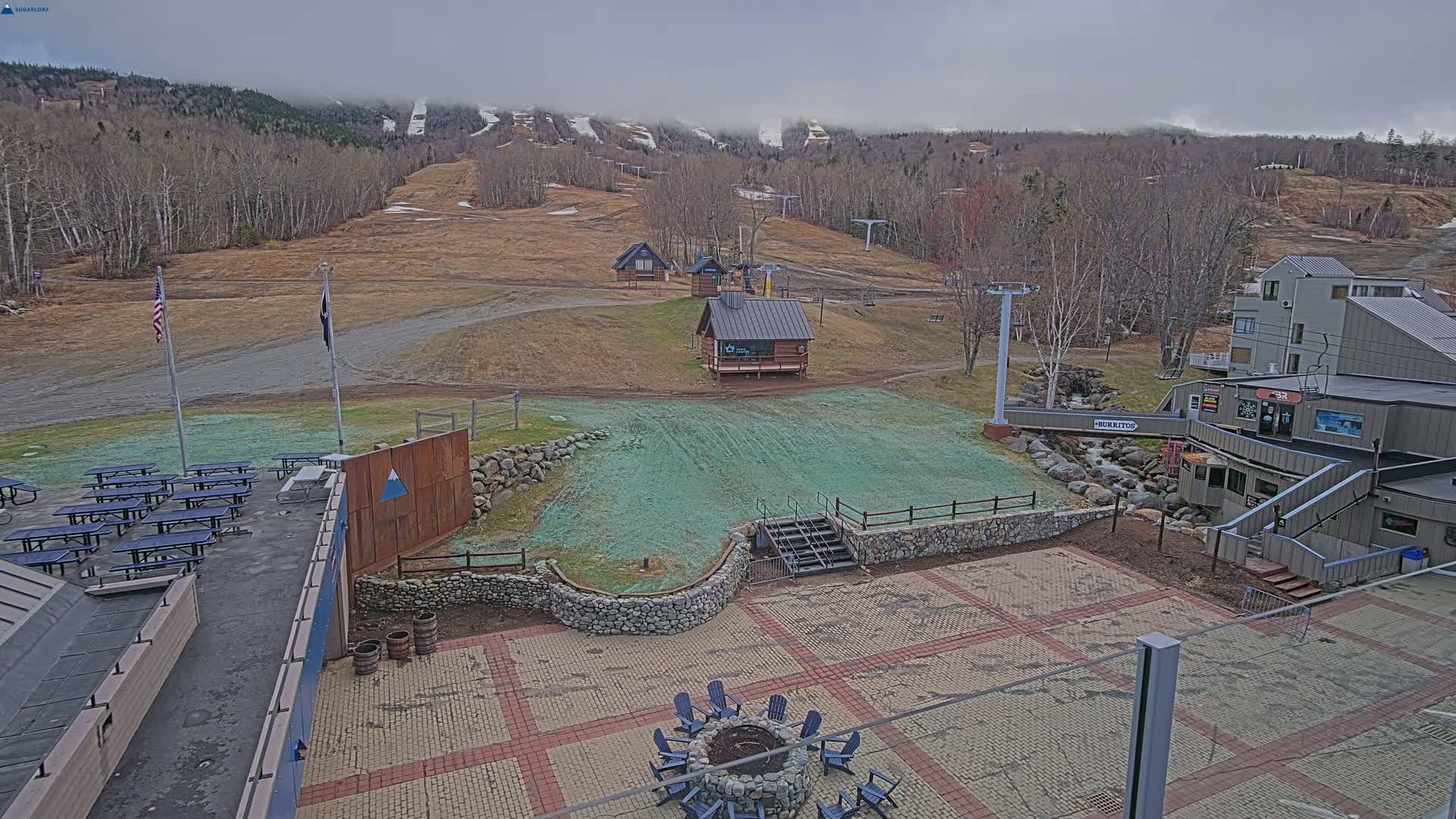 An outdoor patio area with picnic tables and a fire pit overlooks a ski resort with barren slopes and leafless trees under an overcast sky.