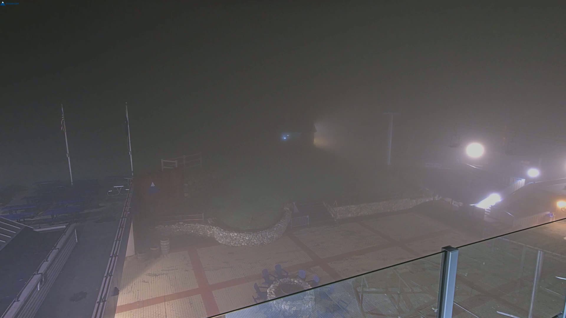 A foggy nighttime scene shows an outdoor patio area with a stone wall, seating, and a fire pit, partially obscured by heavy fog.