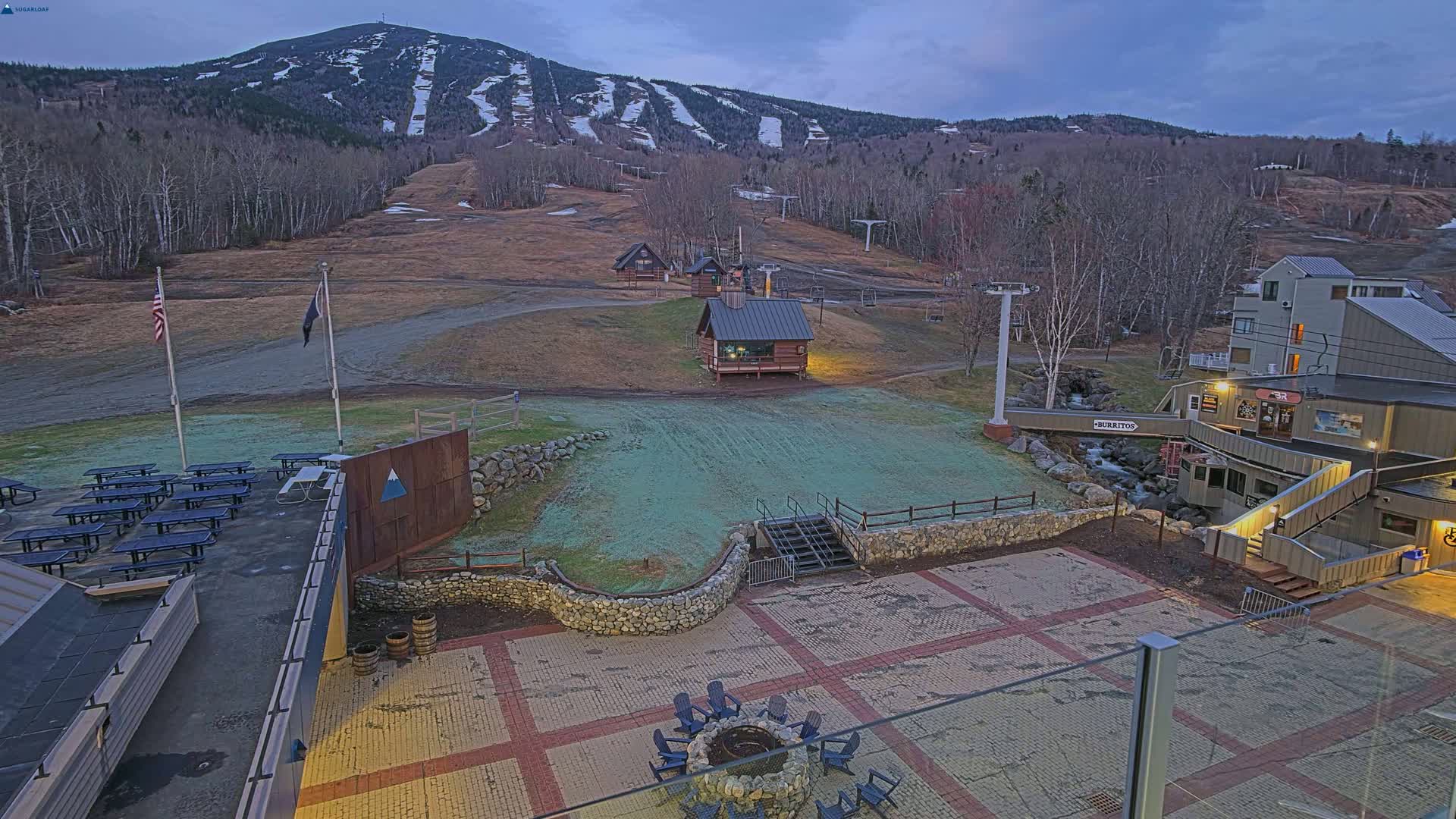 A ski resort at dusk, with a paved area containing picnic tables and a fire pit, overlooks a grassy slope and ski runs on a mountainside.