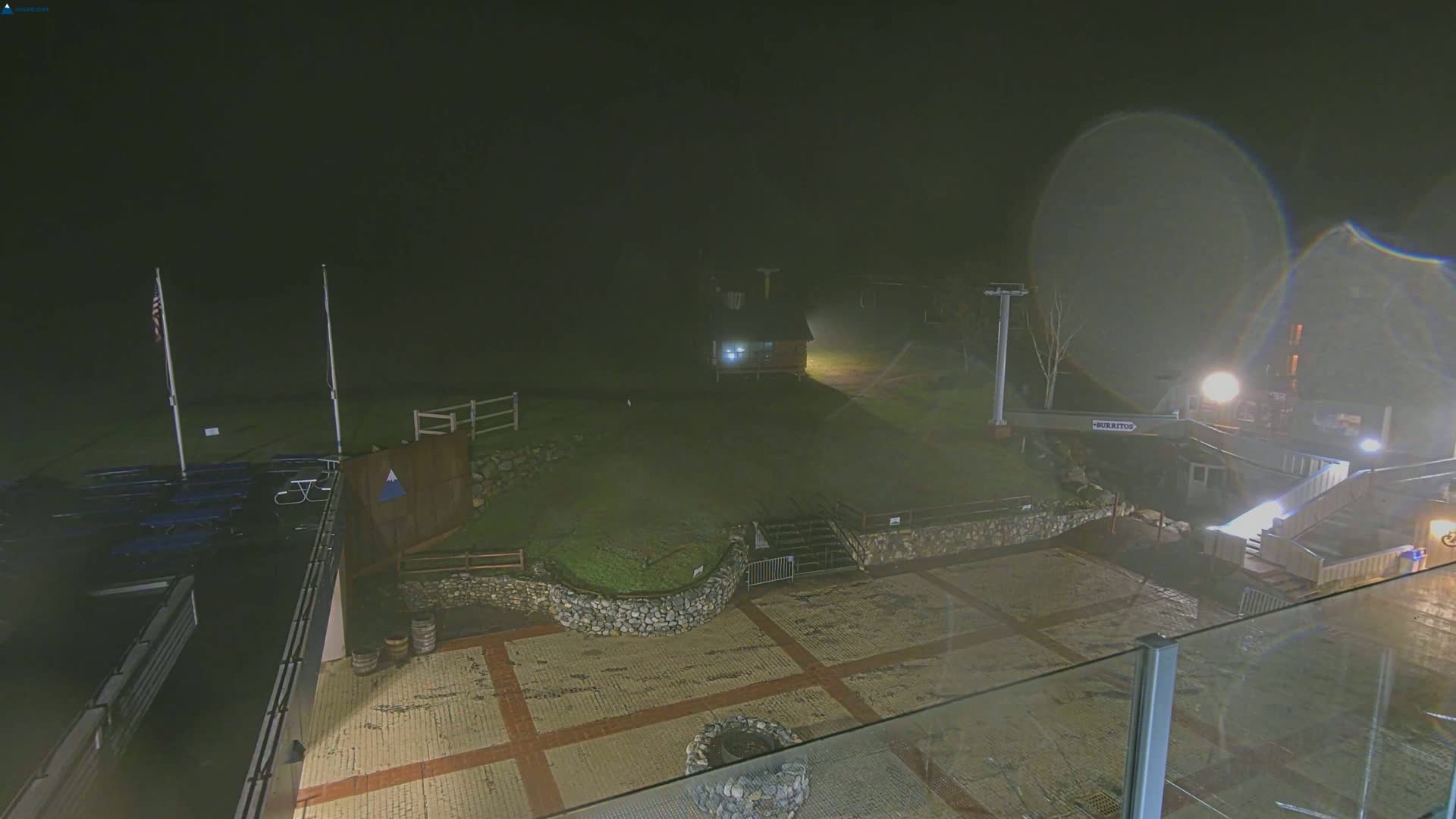 A nighttime, foggy view of an outdoor area with a paved plaza, stone walls, a small building, and ski lift infrastructure.