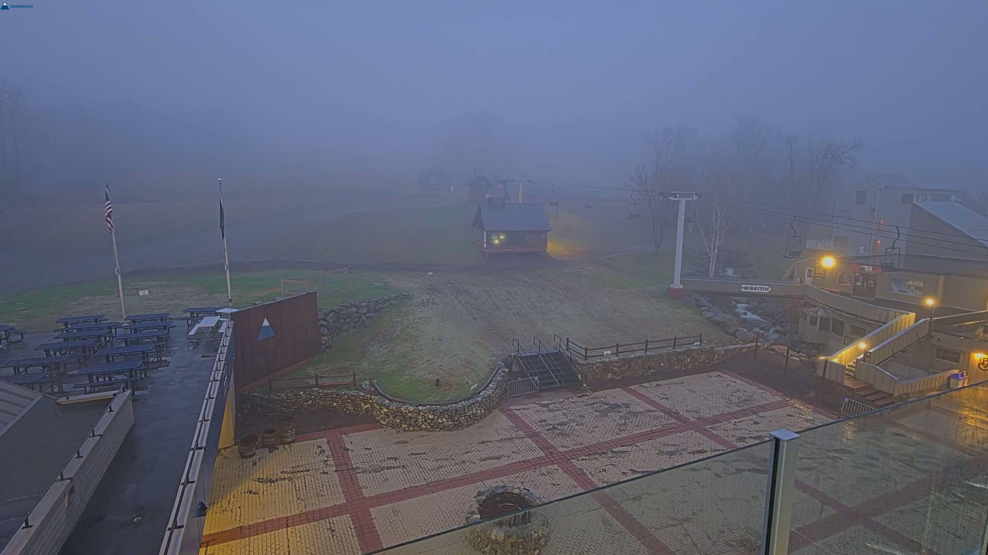 A foggy outdoor scene shows a paved patio area with picnic tables, a small building, a ski lift, and a building in the background.