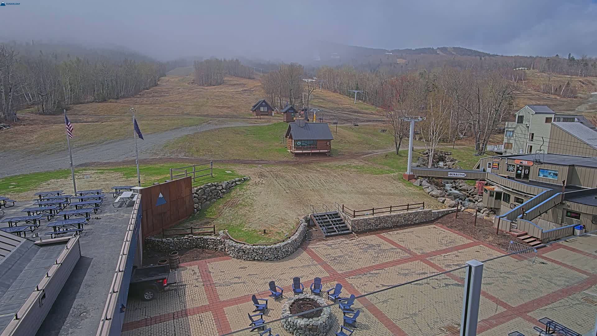 An outdoor patio area with a fire pit and picnic tables overlooks a ski slope with several small buildings under a hazy sky.