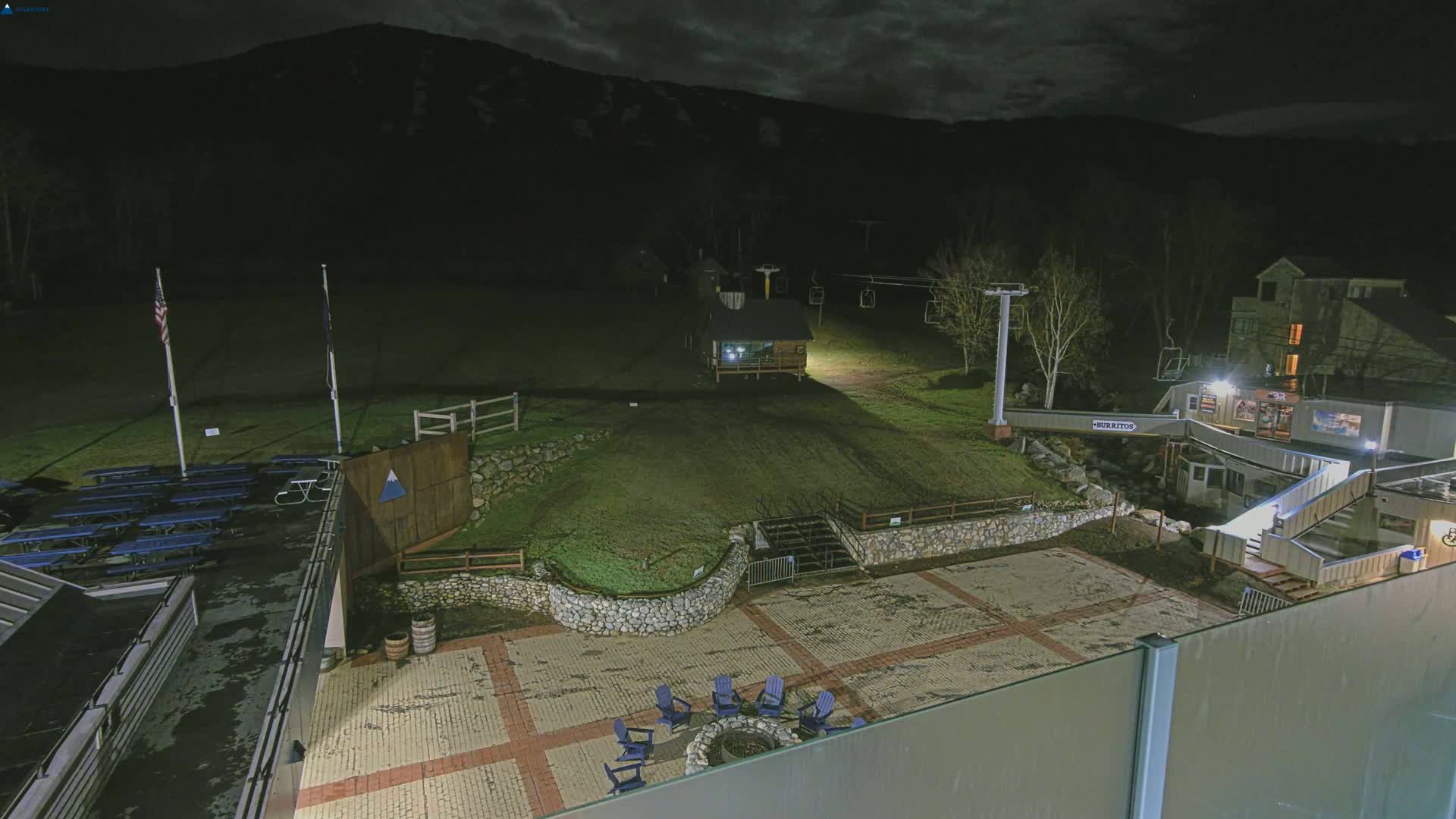A nighttime view of a ski resort base area showing a paved plaza with chairs around a fire pit, a small building, a ski lift, and a dark mountain in the background under a cloudy sky.