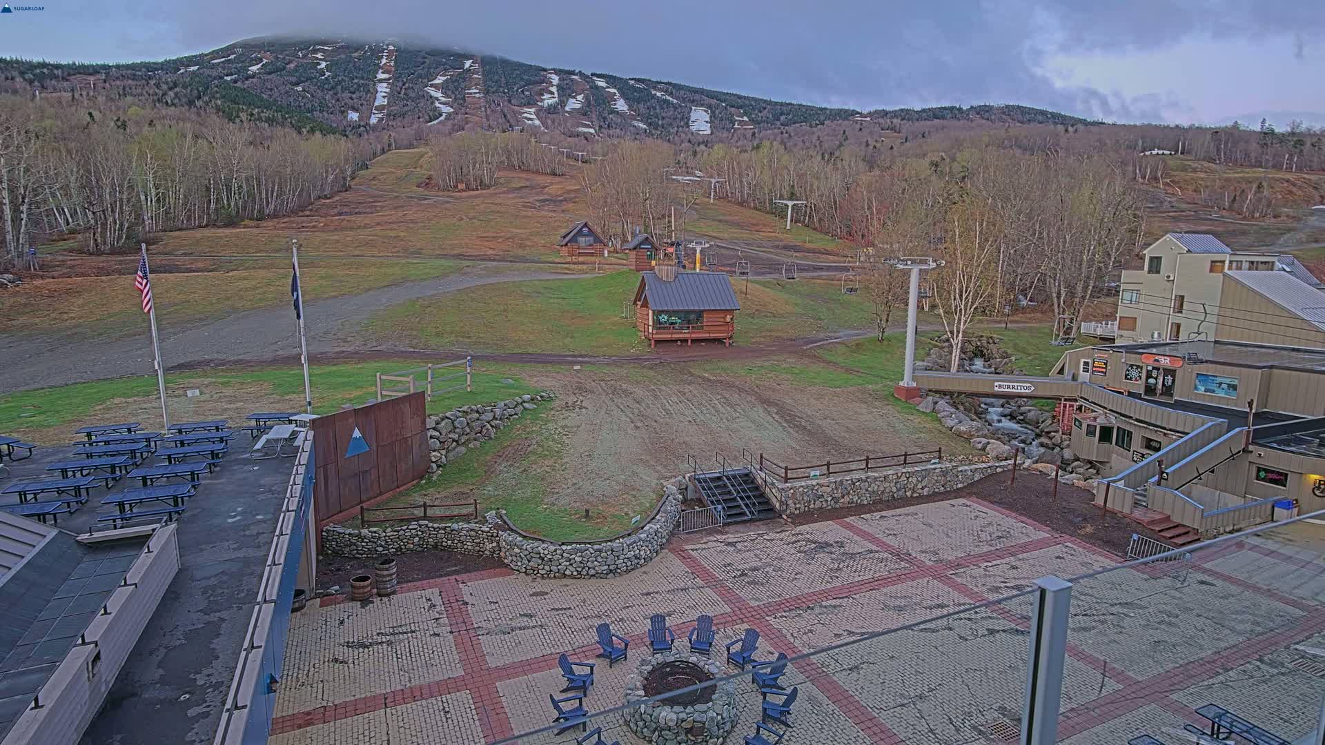 A ski resort base area with a paved plaza, picnic tables, a fire pit, small buildings, and a chairlift, overlooks a mountain with a cloudy sky.